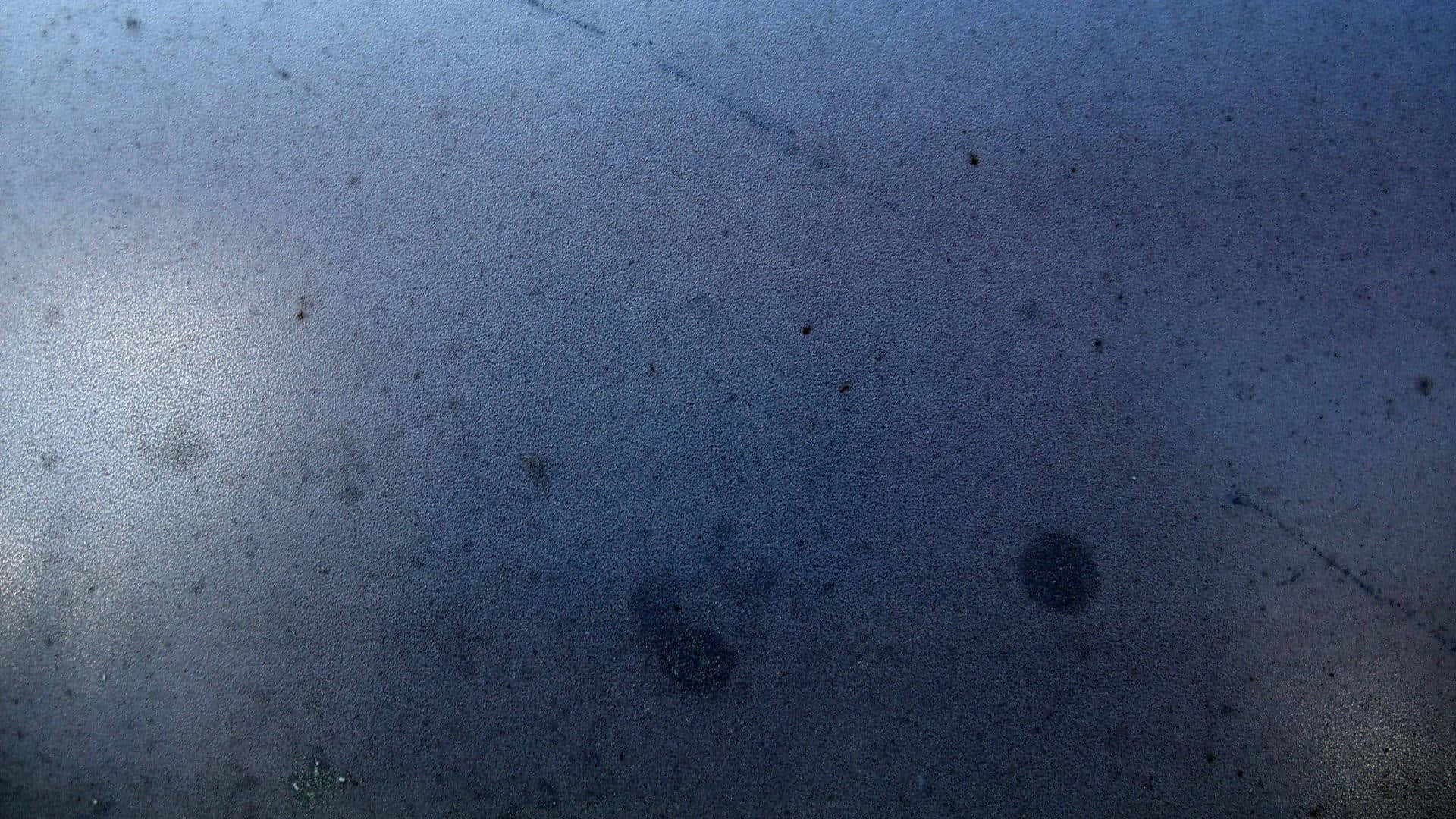 Grey Blue 桌布 壁紙 — 柔和光線下，佈滿灰塵的藍色表面特寫，帶有散落的黑色斑點和輕微刮痕。
