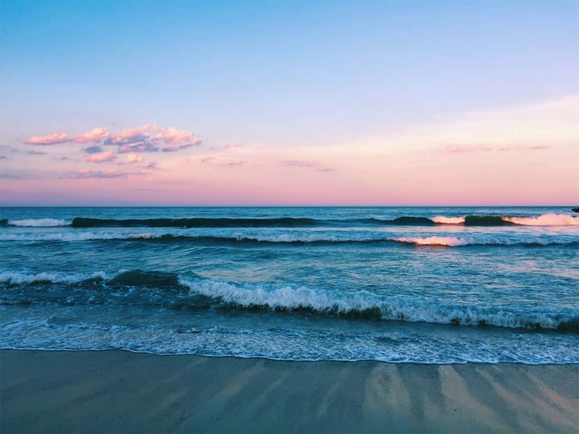 Aesthetic Beach 桌布 壁紙 — 柔和的海浪拍打著沙灘，天空是粉紅和藍色的日落色調，點綴著幾朵雲彩。