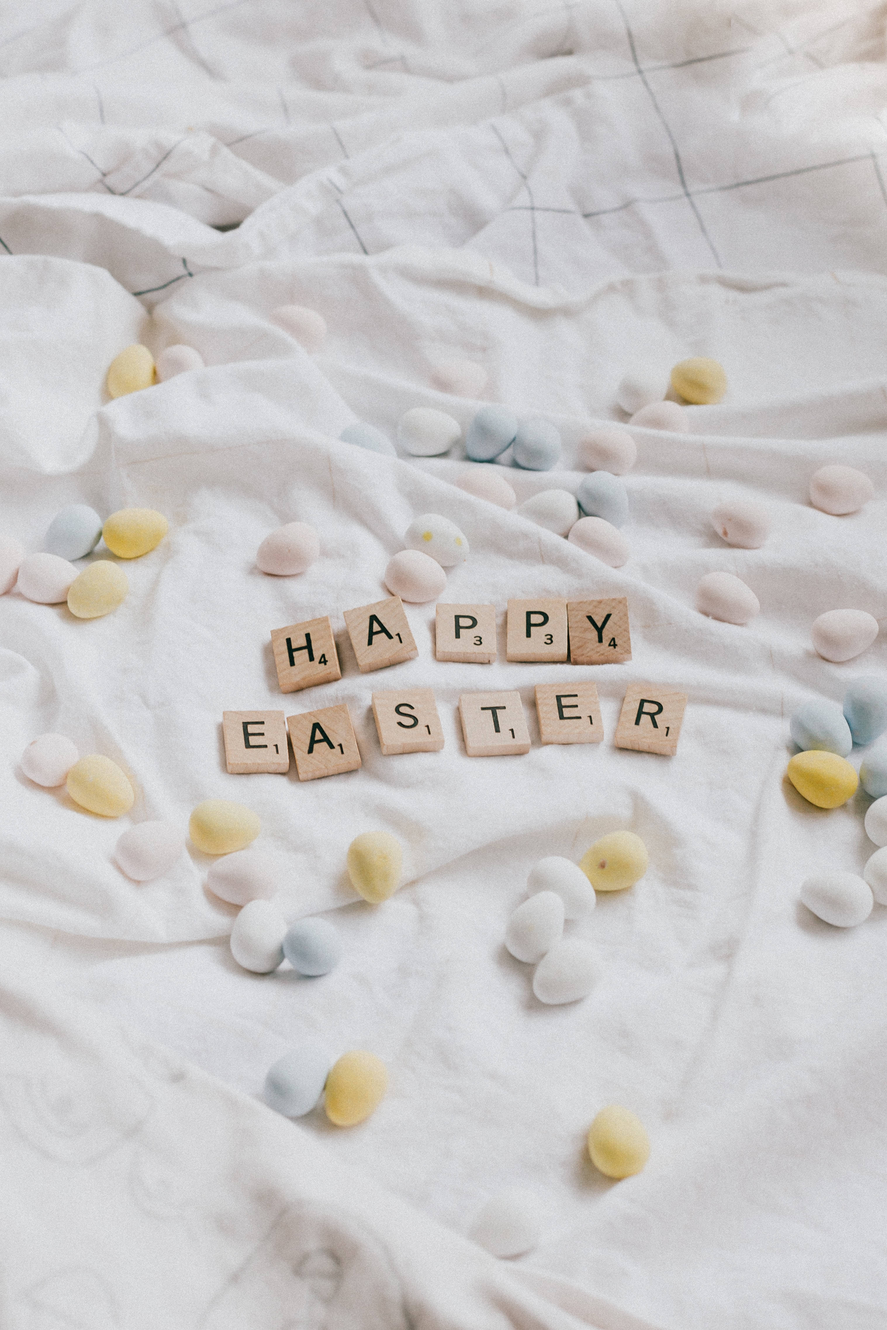 Easter Phone 桌布 壁紙 — 柔和的粉彩復活節蛋和木製Scrabble字母牌拼出'HAPPY EASTER'，放在皺巴巴的白色布料上。
