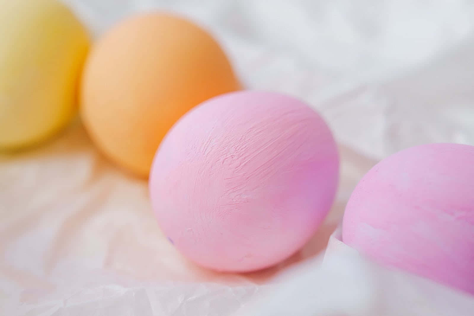 Pastel Easter 桌布 壁紙 — 柔和的粉彩蛋在黃色、橙色和粉紅色中，放在皺巴巴的白色布料上，景深較淺。