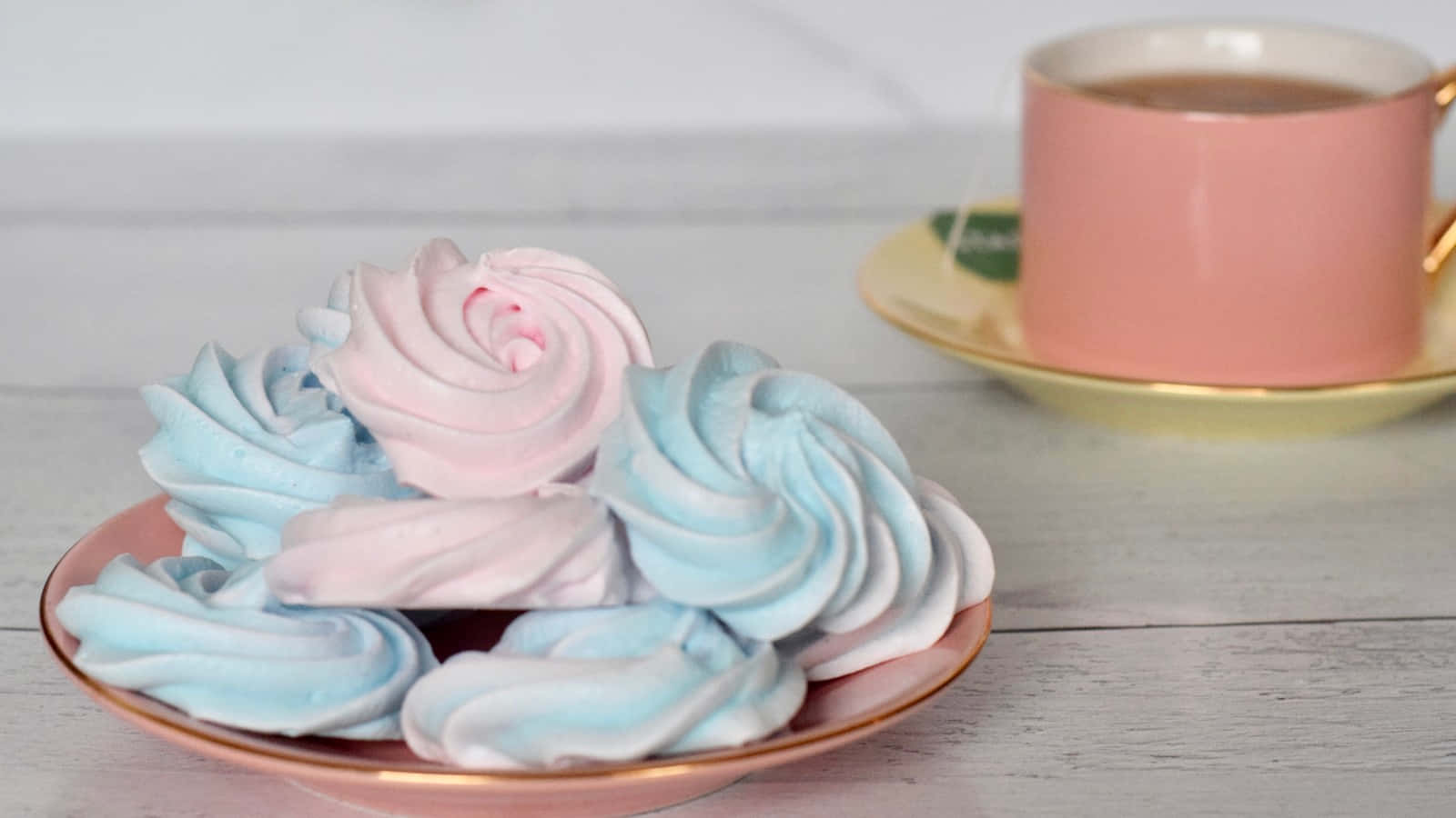 Meringue 桌布 壁紙 — 柔和的粉藍色和粉紅色蛋白霜玫瑰優雅地放在玫瑰金盤子上，旁邊有一個粉紅色茶杯，置於白色木桌上。