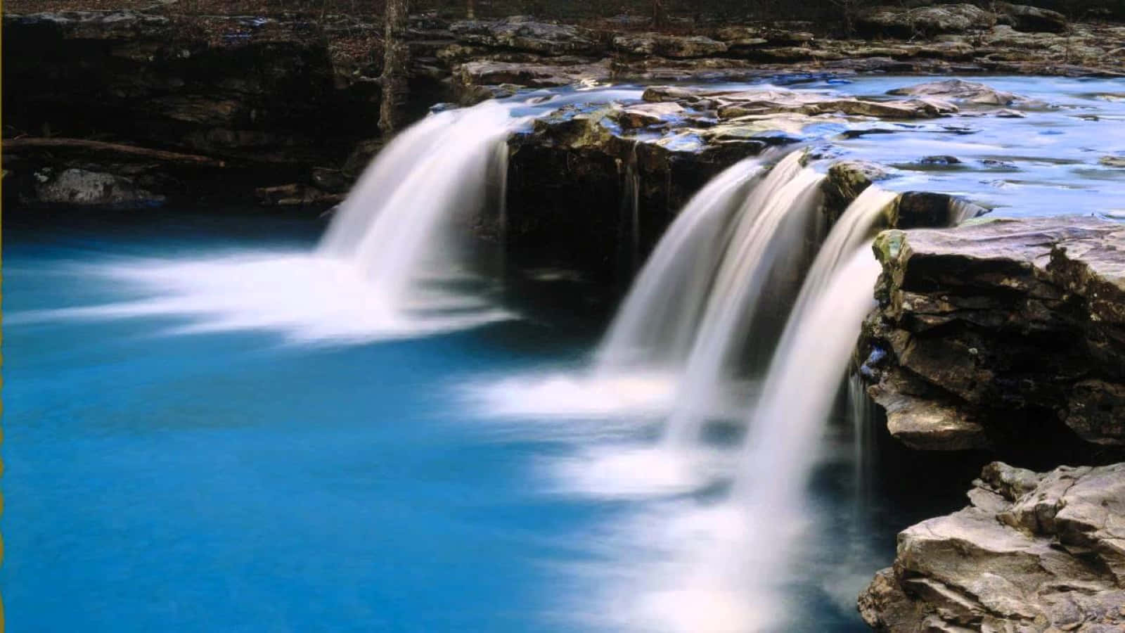 3d Waterfall 桌布 壁紙 — 柔和的自然光下，寧靜的瀑布從深色岩石上傾瀉而下，流入充滿活力的藍色水池。