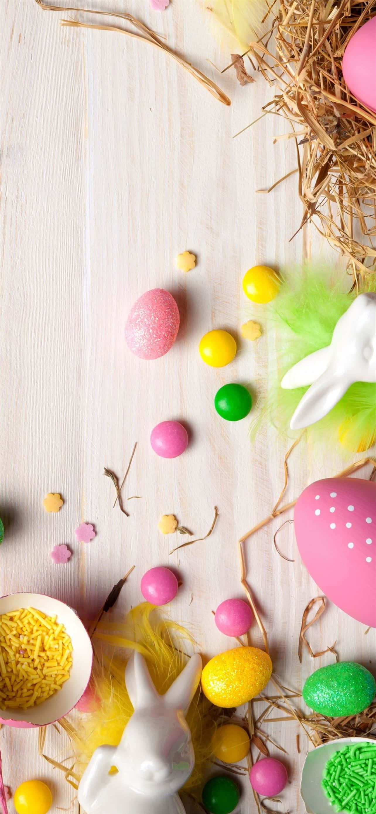 Cute Easter iPhone 桌布 壁紙 — 柔和色調的復活節彩蛋和兔子公仔散落在淺色木質表面上，伴有乾草和彩色糖粒。