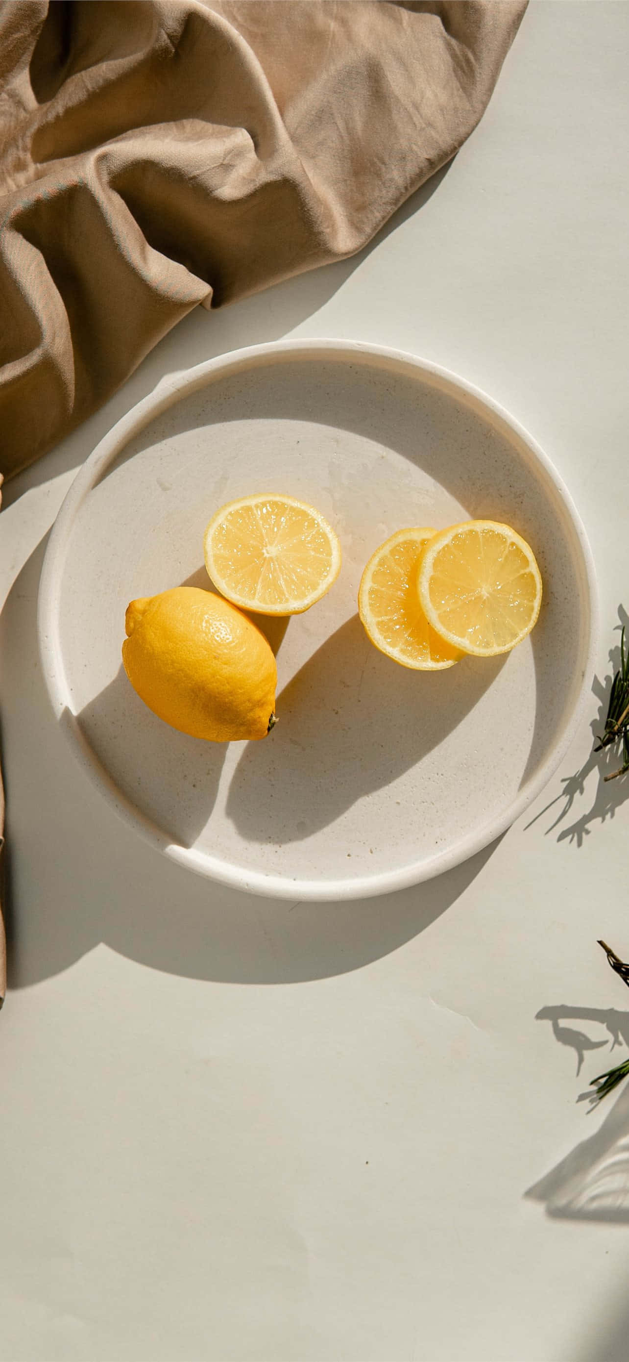 Lemon iPhone 桌布 壁紙 — 柔和陽光下，一個有斑點的白色盤子上放著鮮黃色檸檬，旁邊有米色布料和迷迭香枝條的俯視圖。