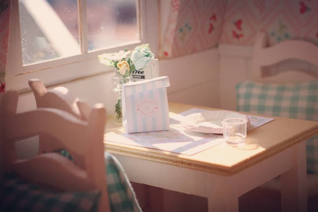 娃娃屋 桌布 壁紙 — 柔和陽光灑在木桌上，桌上放有條紋盒子花朵和玻璃杯，旁邊是花卉壁紙的窗戶。