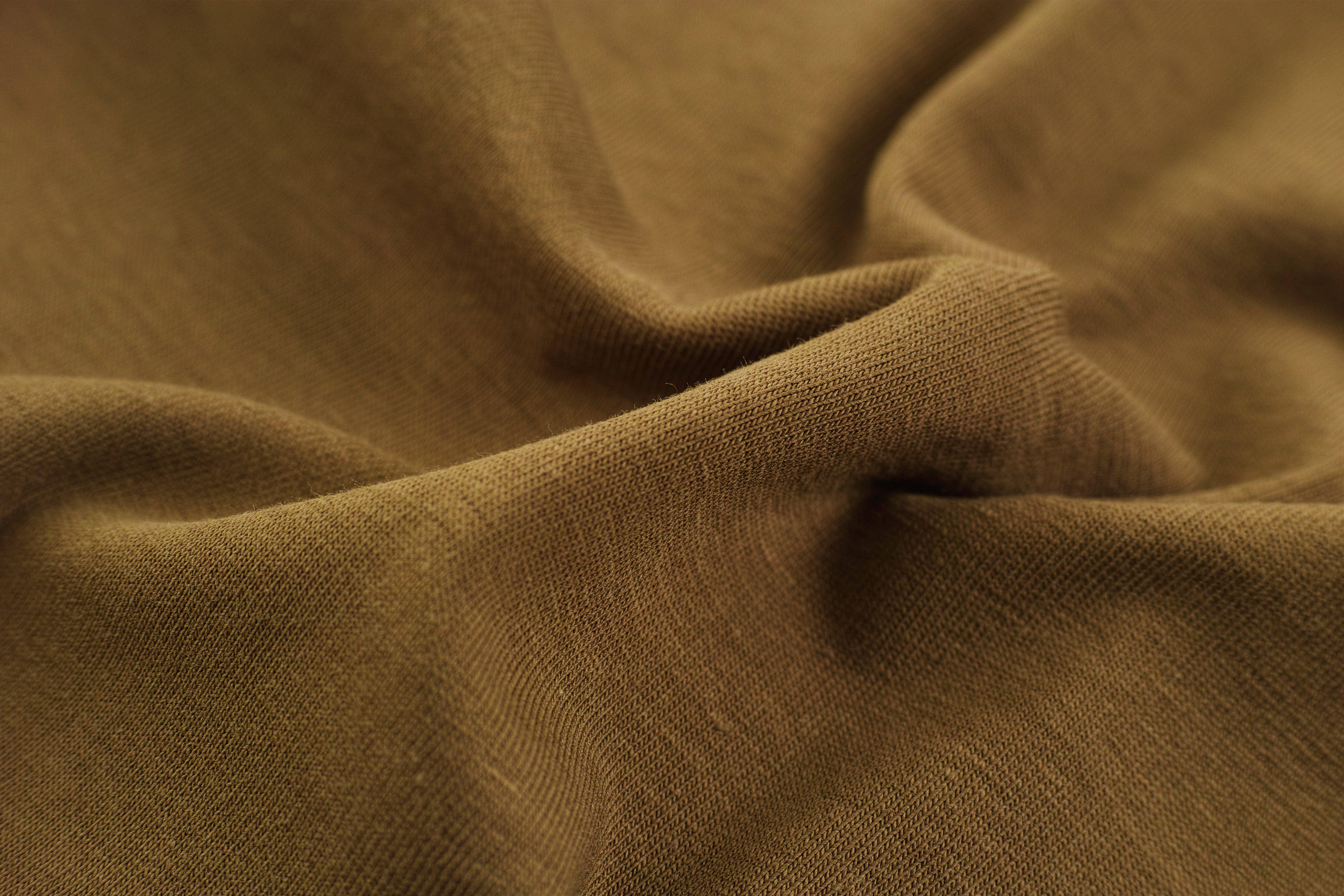 Textured 桌布 壁紙 — 柔軟棕色布料的特寫，在溫暖燈光下呈現柔和摺痕和可見的編織紋理。