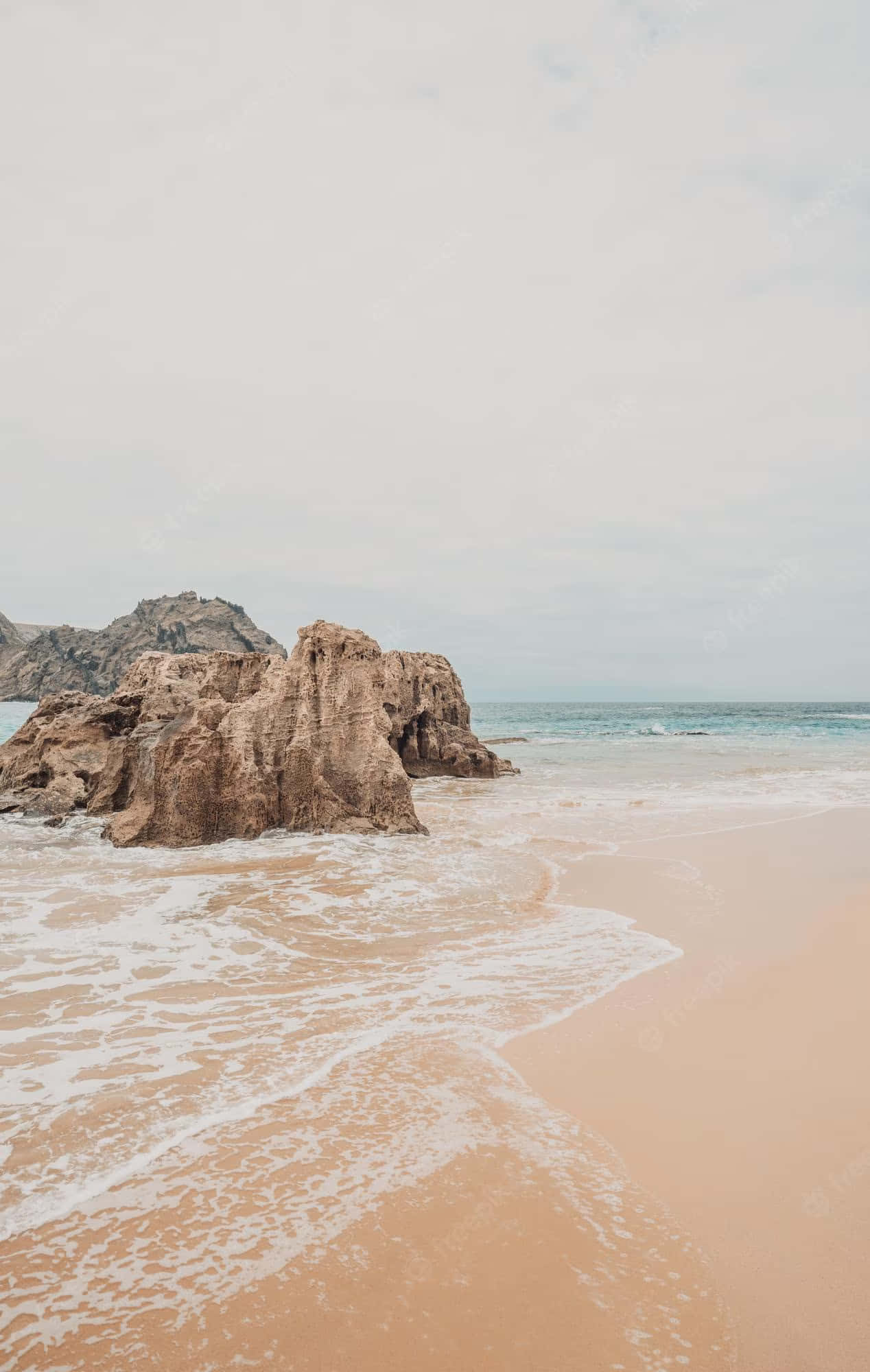 Beach Aesthetic 桌布 壁紙 — 柔軟的波浪拍打著淡粉紅色的沙灘，旁邊是風化的棕色海蝕柱，天空多雲。