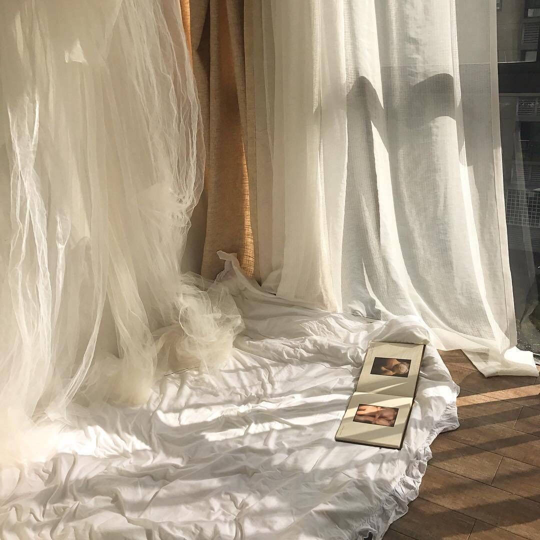 Tan 桌布 壁紙 — 柔軟的白色窗簾垂落在陽光照耀的木地板上，一本相簿放在皺巴巴的床單上。