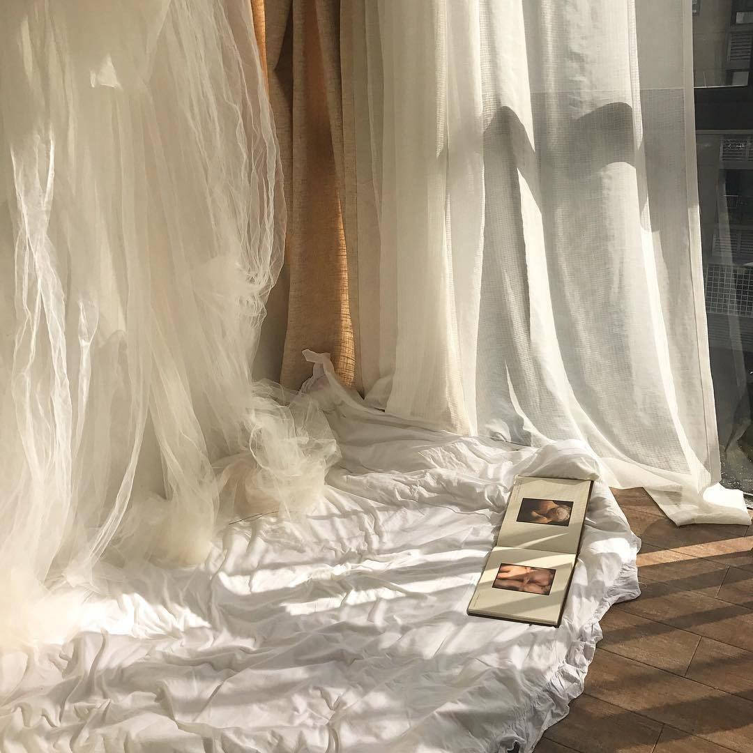 美學米色 桌布 壁紙 — 柔軟的白色窗簾垂落在陽光照耀的木地板上，上面放著一張皺巴巴的白色床單和一本打開的相簿。