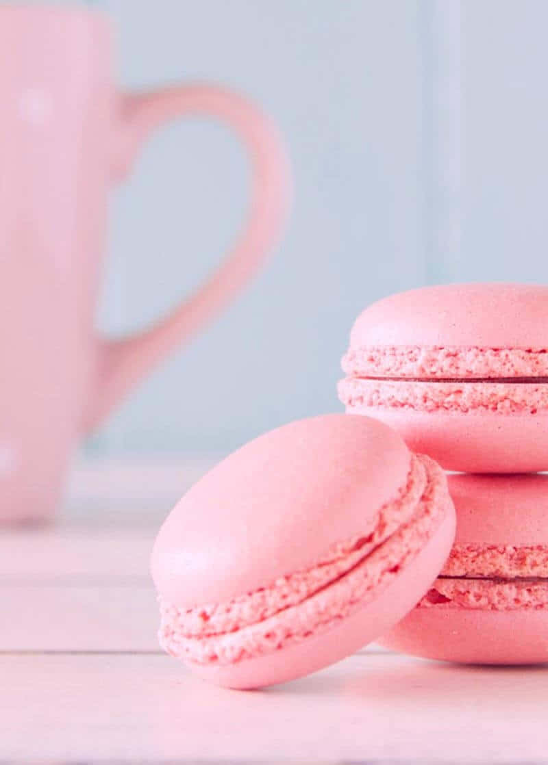 Macarons 桌布 壁紙 — 柔軟的粉紅色馬卡龍放在木質表面上，旁邊有一個模糊的粉紅色杯子，背景是淺藍色牆壁。