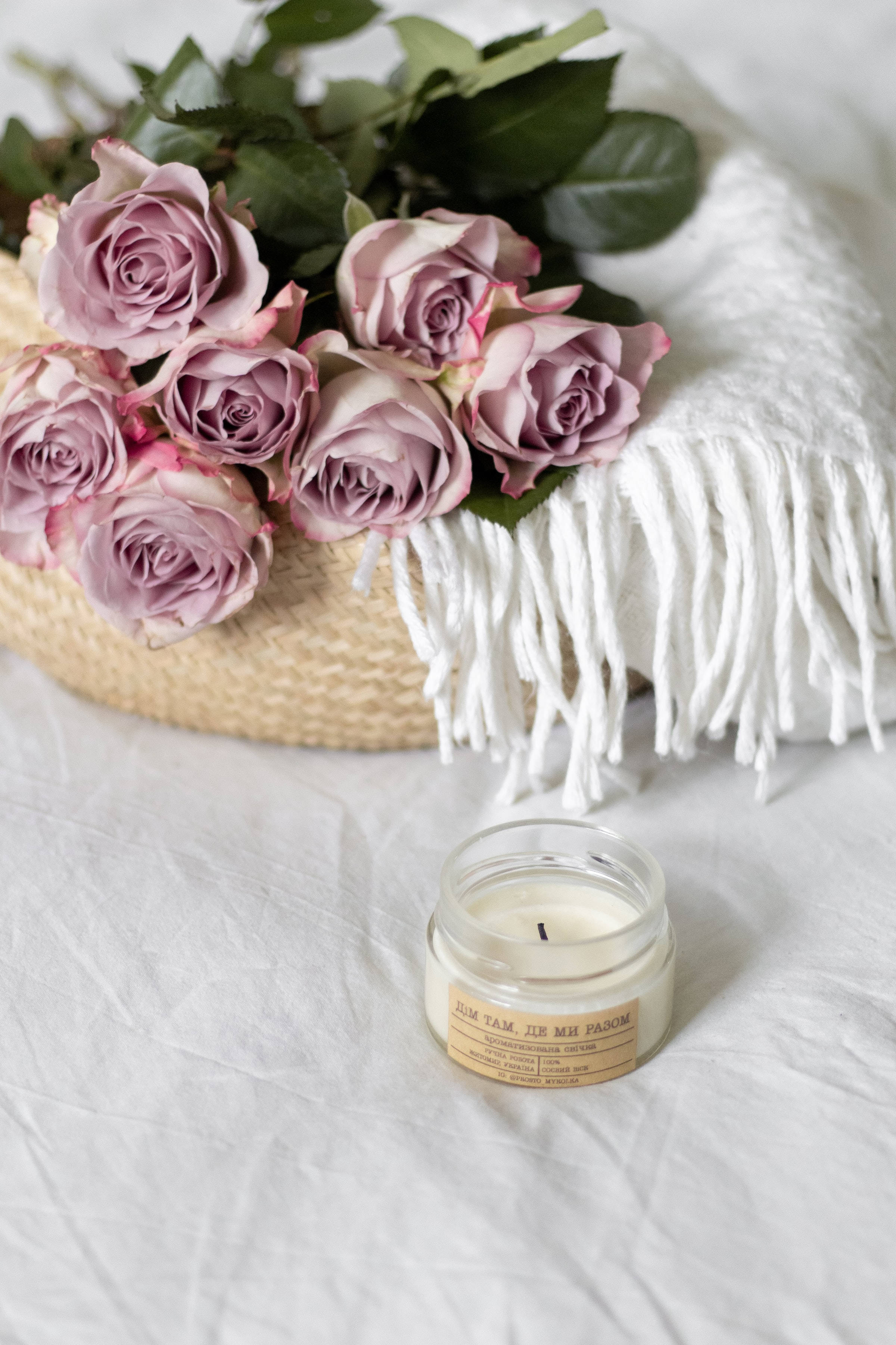 Roses 桌布 壁紙 — 柔軟的紫紅色玫瑰放在編織籃中，旁邊有一條白色流蘇毯子和一個玻璃蠟燭罐，放在皺巴巴的白色布料上。