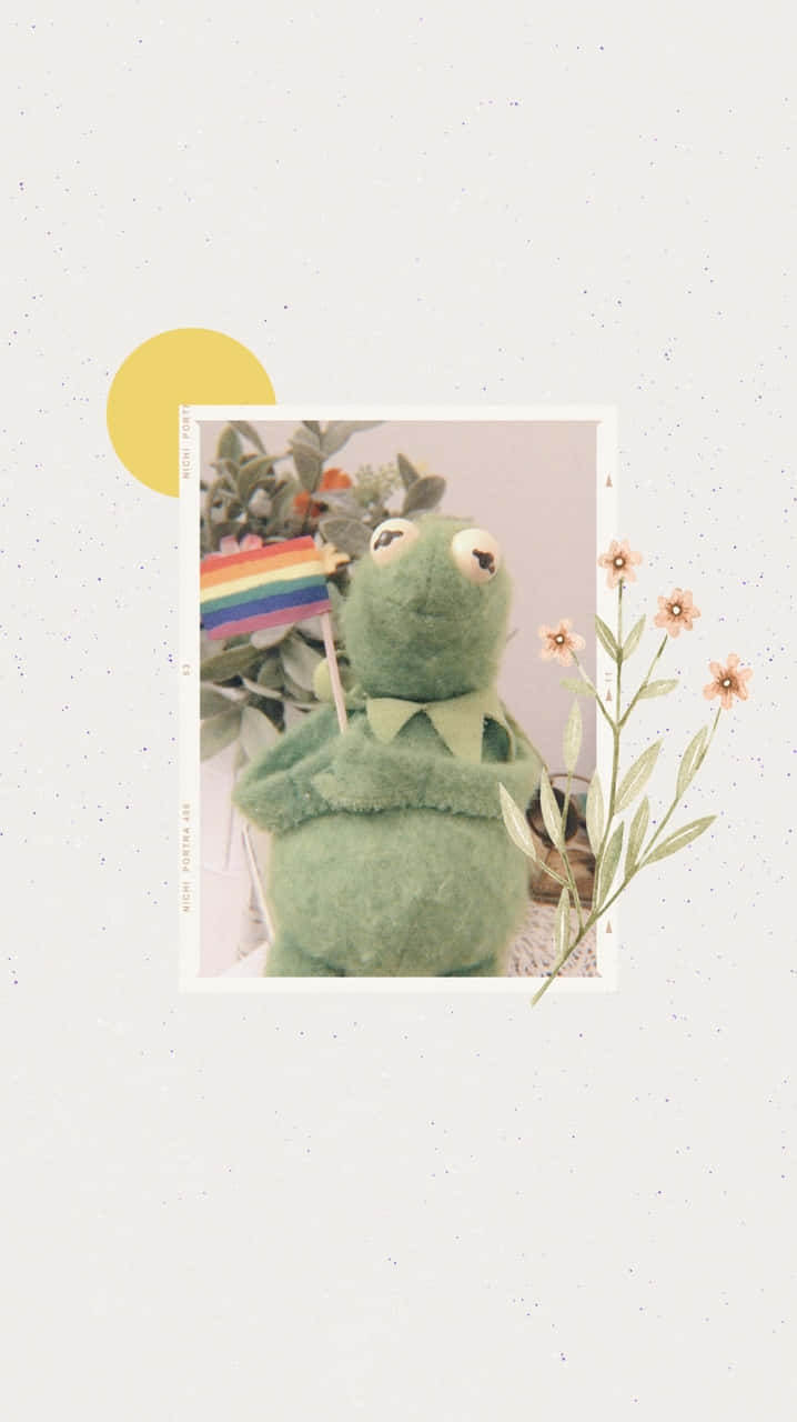 Aesthetic Kermit 桌布 壁紙 — 柔軟的綠色青蛙布偶在精緻的花朵和黃色太陽中舉起彩虹旗，背景帶有斑點。
