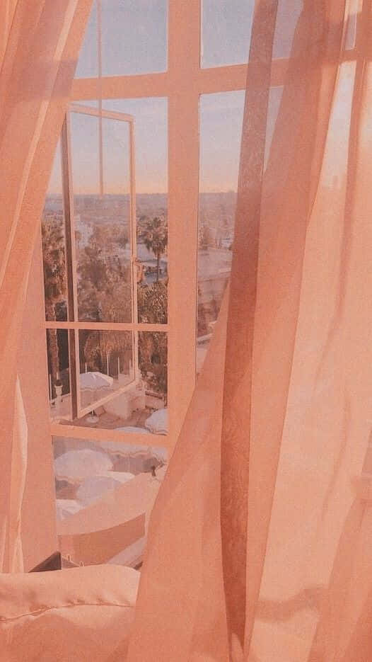 Peachy Aesthetic 桌布 壁紙 — 柔軟的蜜桃色窗簾框住一扇窗，窗外是陽光普照的城市景觀，棕櫚樹和屋頂沐浴在溫暖的金色光芒中。