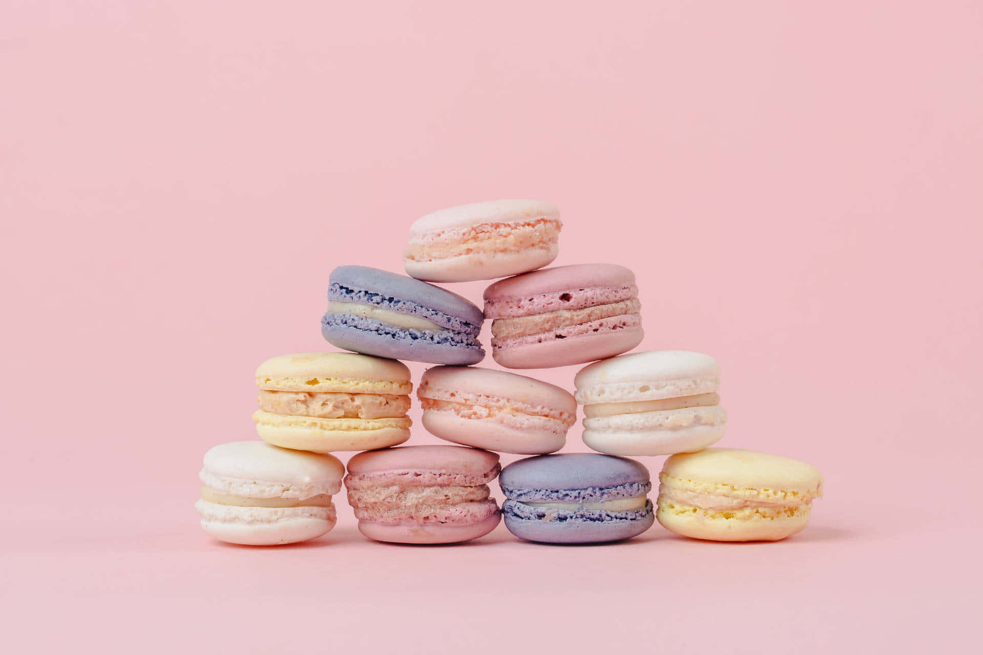 Macarons 桌布 壁紙 — 柔軟粉紅、黃色和薰衣草色調的粉彩馬卡龍堆疊成金字塔，背景為純色粉紅。