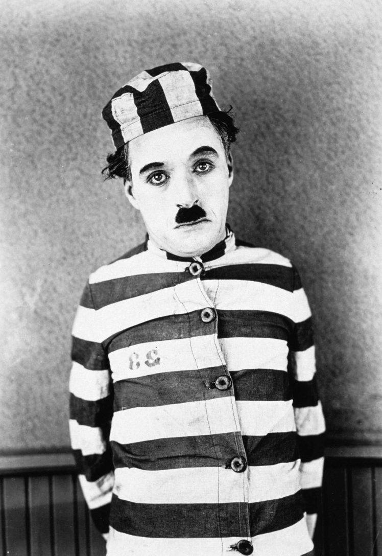 Chaplin 桌布 壁紙 — 查理·卓別林穿著標誌性的監獄制服，條紋襯衫和帽子，用富有表現力的眼睛和鬍子直視鏡頭。
