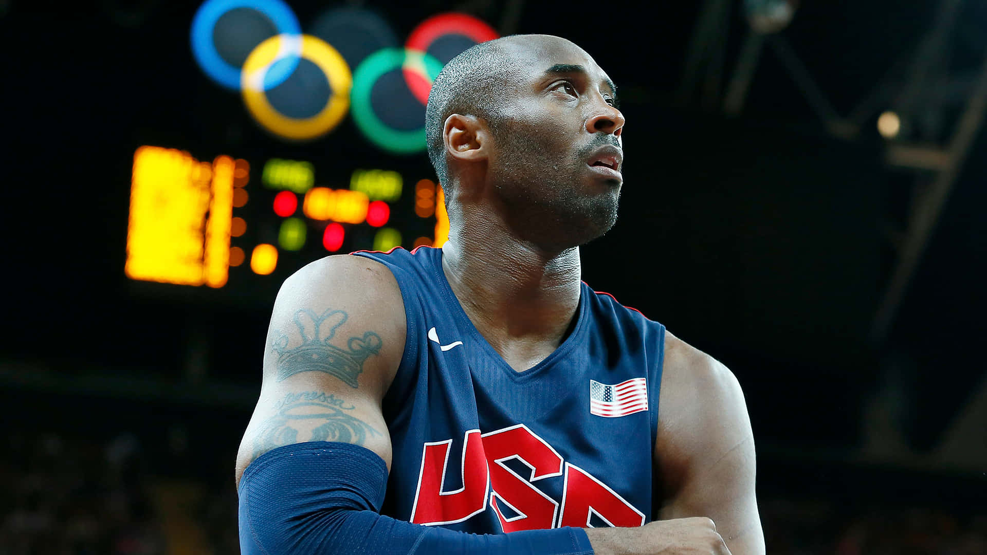 Mamba Mentality 桌布 壁紙 — 柯比·布萊恩穿著美國籃球球衣，在場館燈光下專注地向上凝視，身後奧運五環閃耀。