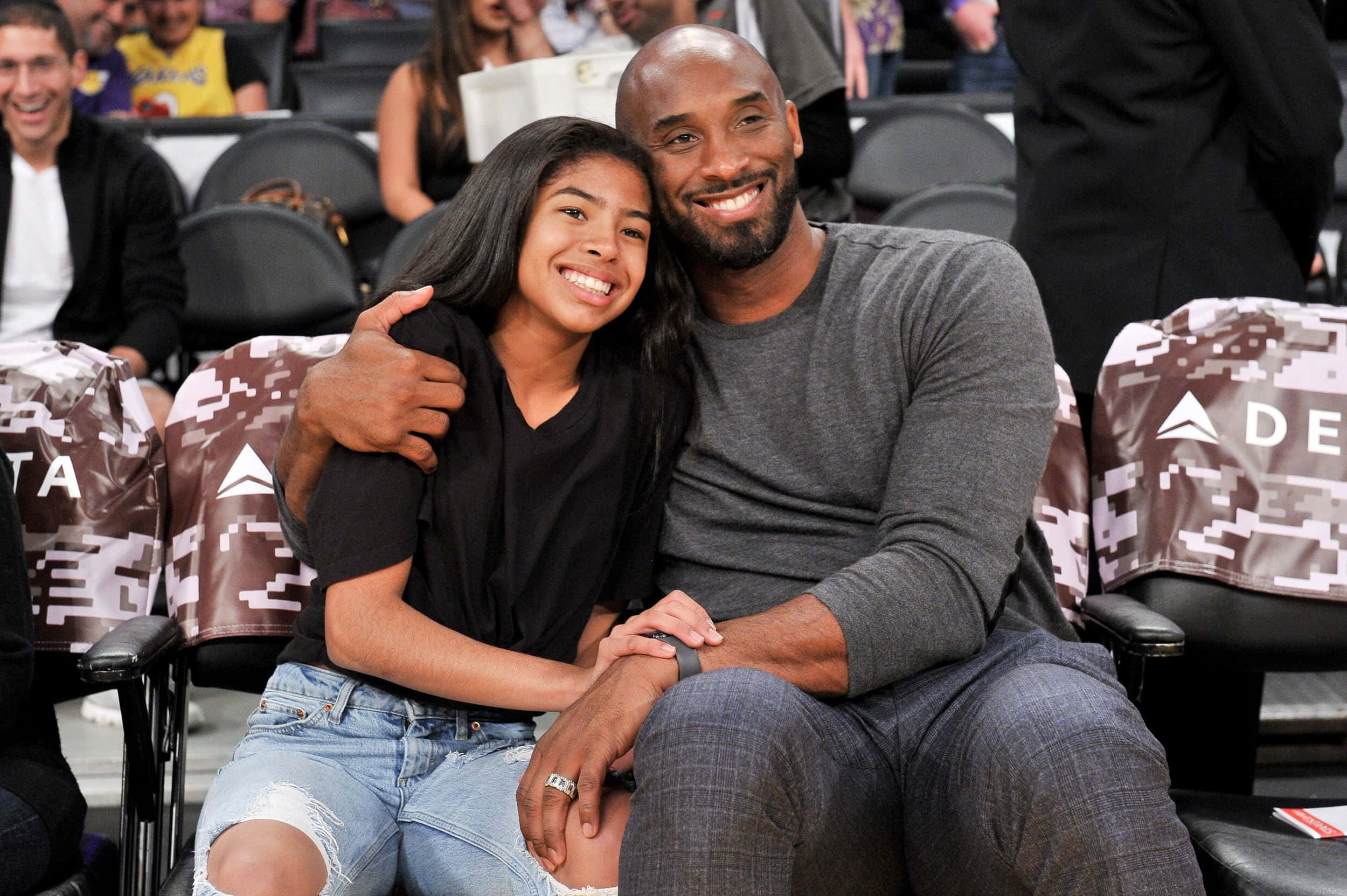 Kobe And Gigi 桌布 壁紙 — 柯比·布萊恩與女兒吉安娜在籃球比賽中坐在一起，背景是體育館座位和球迷，溫暖地微笑。