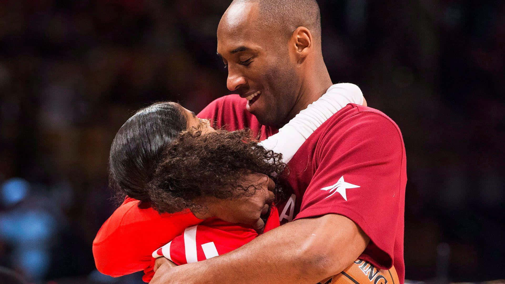 Kobe And Gigi 桌布 壁紙 — 柯比布萊恩在籃球活動中，以溫暖的笑容擁抱一位身穿紅色球衣的小女孩。