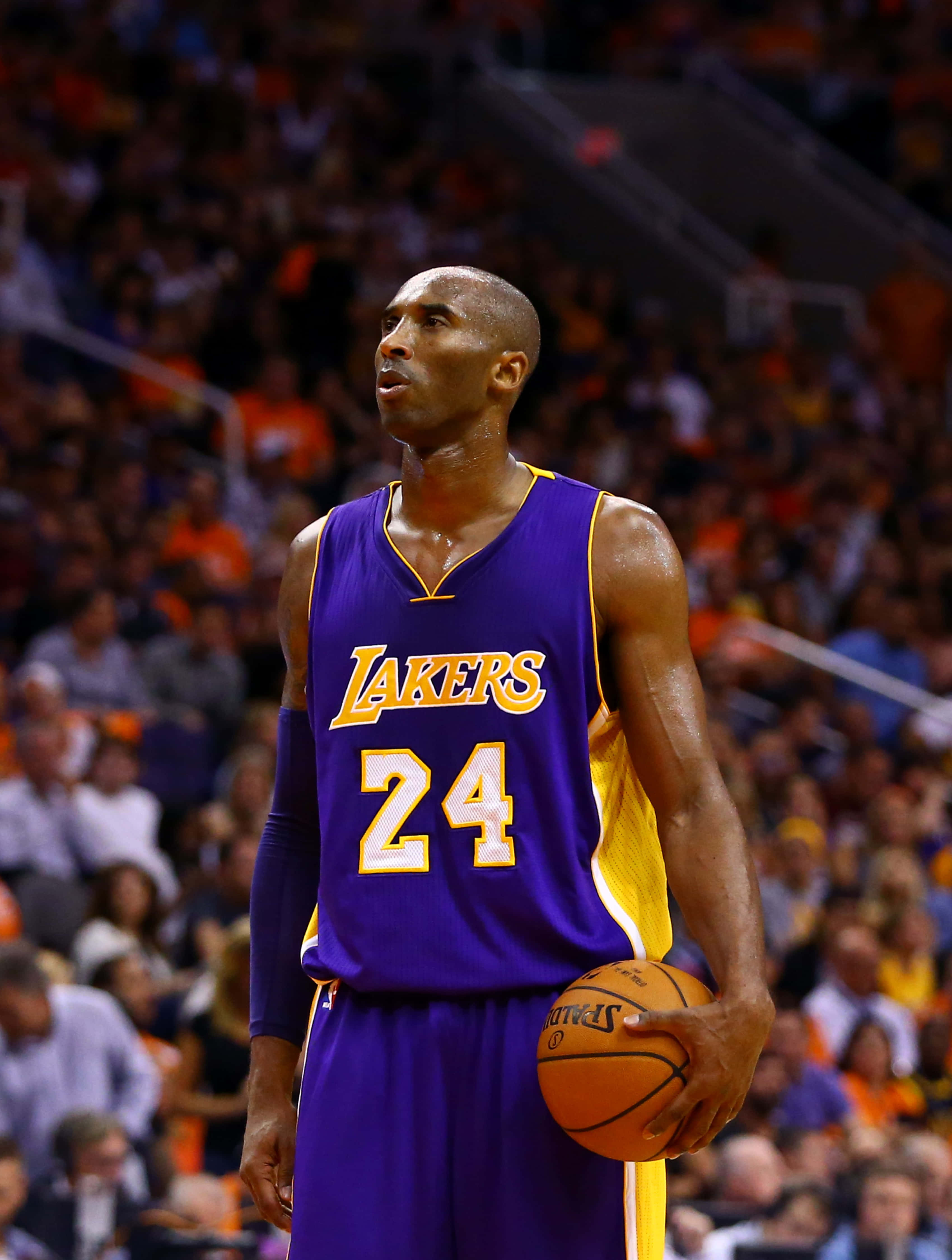 Kobe Basketball 桌布 壁紙 — 柯比布萊恩身穿紫色湖人隊24號球衣，在比賽中專注地拿著籃球，背景是模糊的人群。