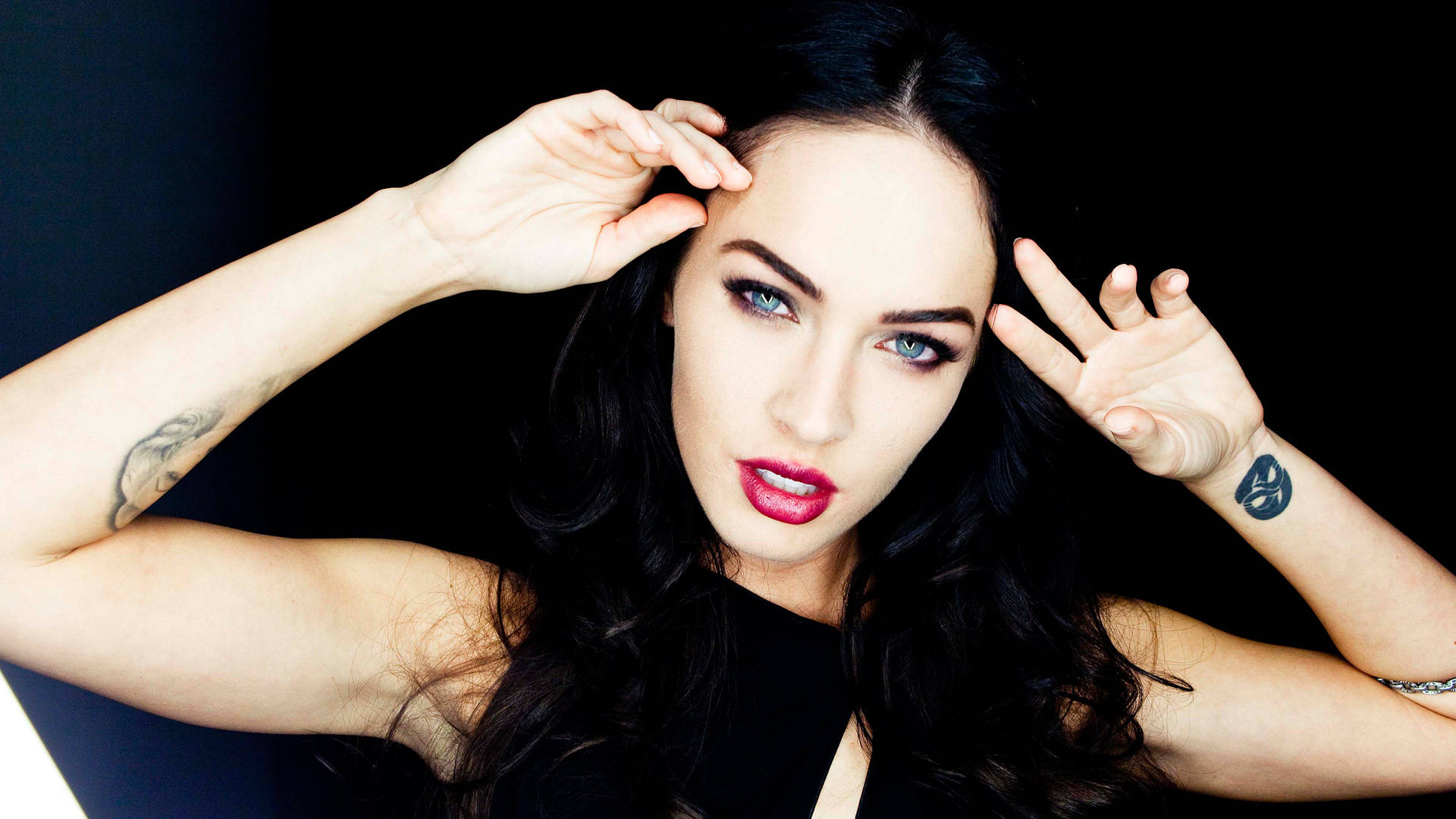 Megan Fox HD 桌布 壁紙 — 梅根·福克斯在純黑背景下將雙手靠近頭部擺姿勢，展現大膽的紅唇和刺青。