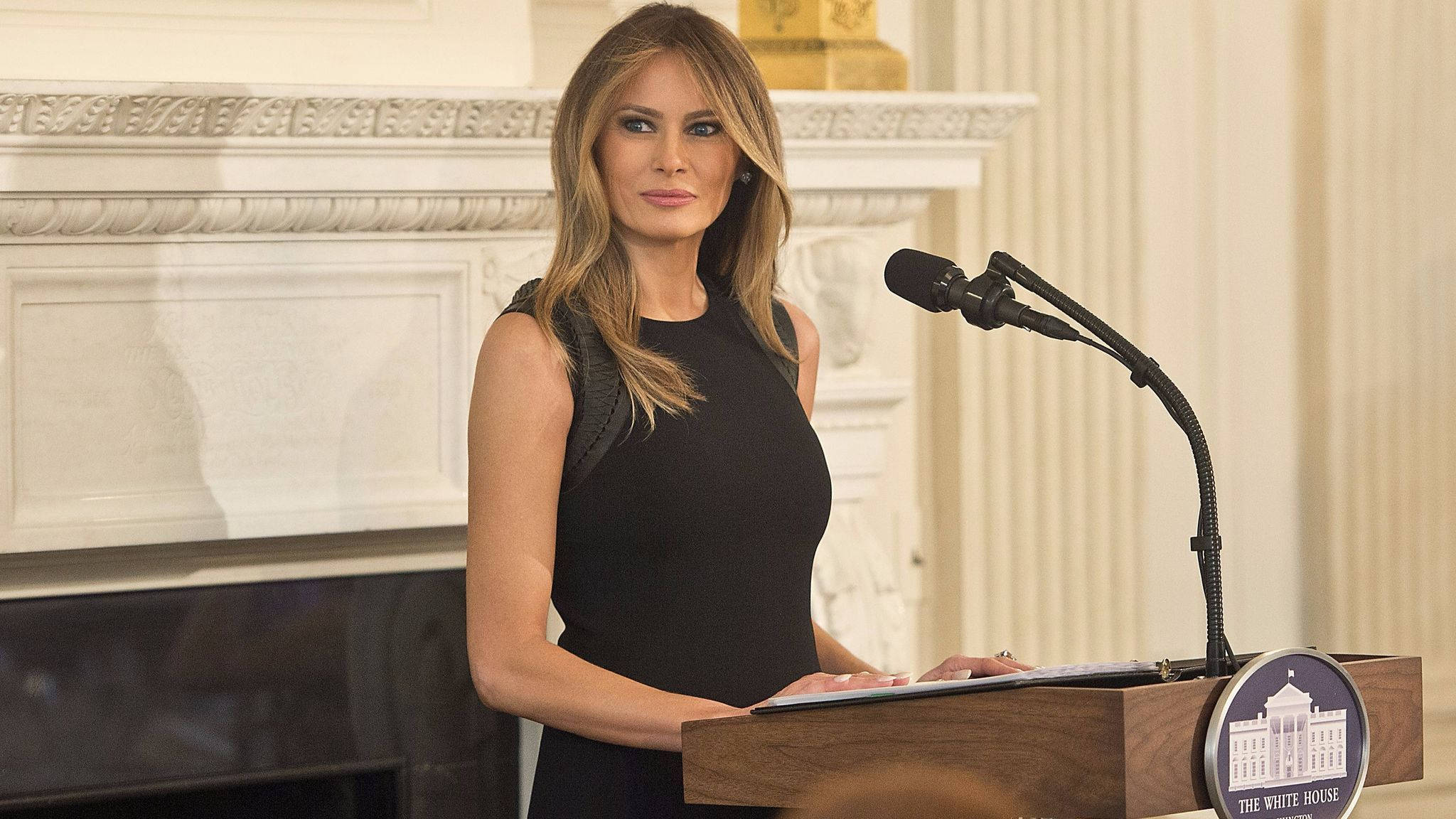 Melania 桌布 壁紙 — 梅蘭妮亞·川普在白宮講台上站穩，穿著正式黑色連衣裙發表演講，身後有華麗的壁爐。