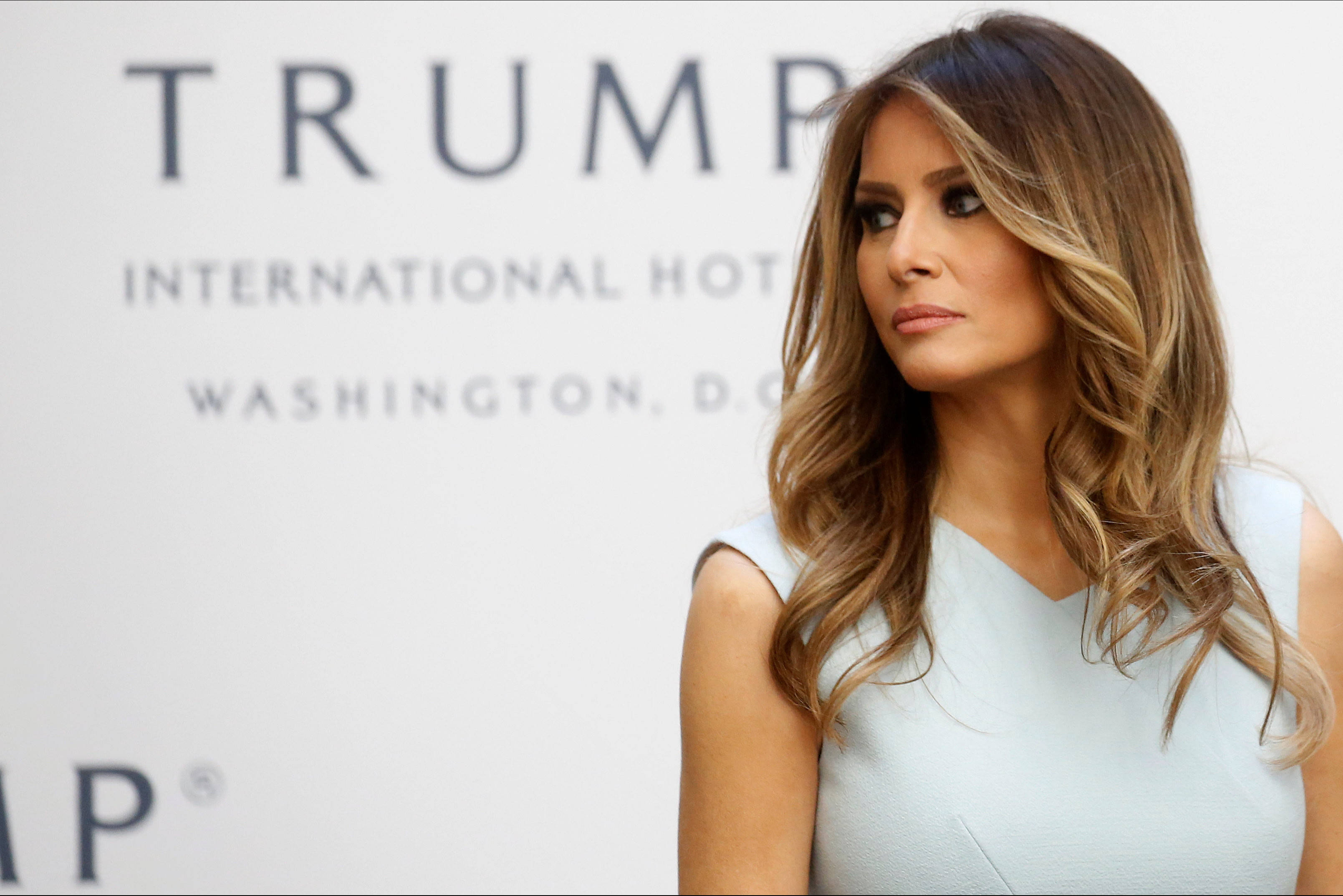 Melania 桌布 壁紙 — 梅蘭妮亞·川普站在華盛頓特區的川普國際飯店標誌前，身穿淺藍色無袖連衣裙，棕色波浪髮。