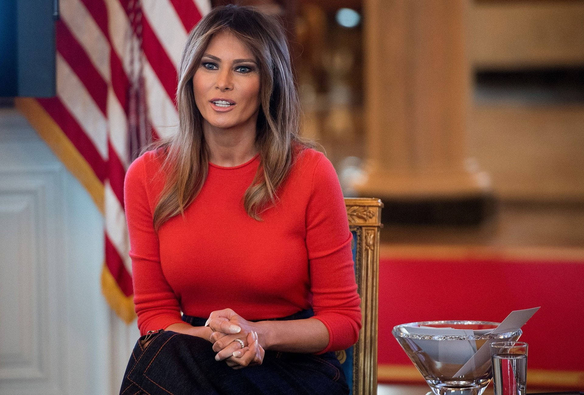 Melania 桌布 壁紙 — 梅蘭妮亞·川普身穿紅色上衣，在美國國旗與華麗木雕背景下端坐。