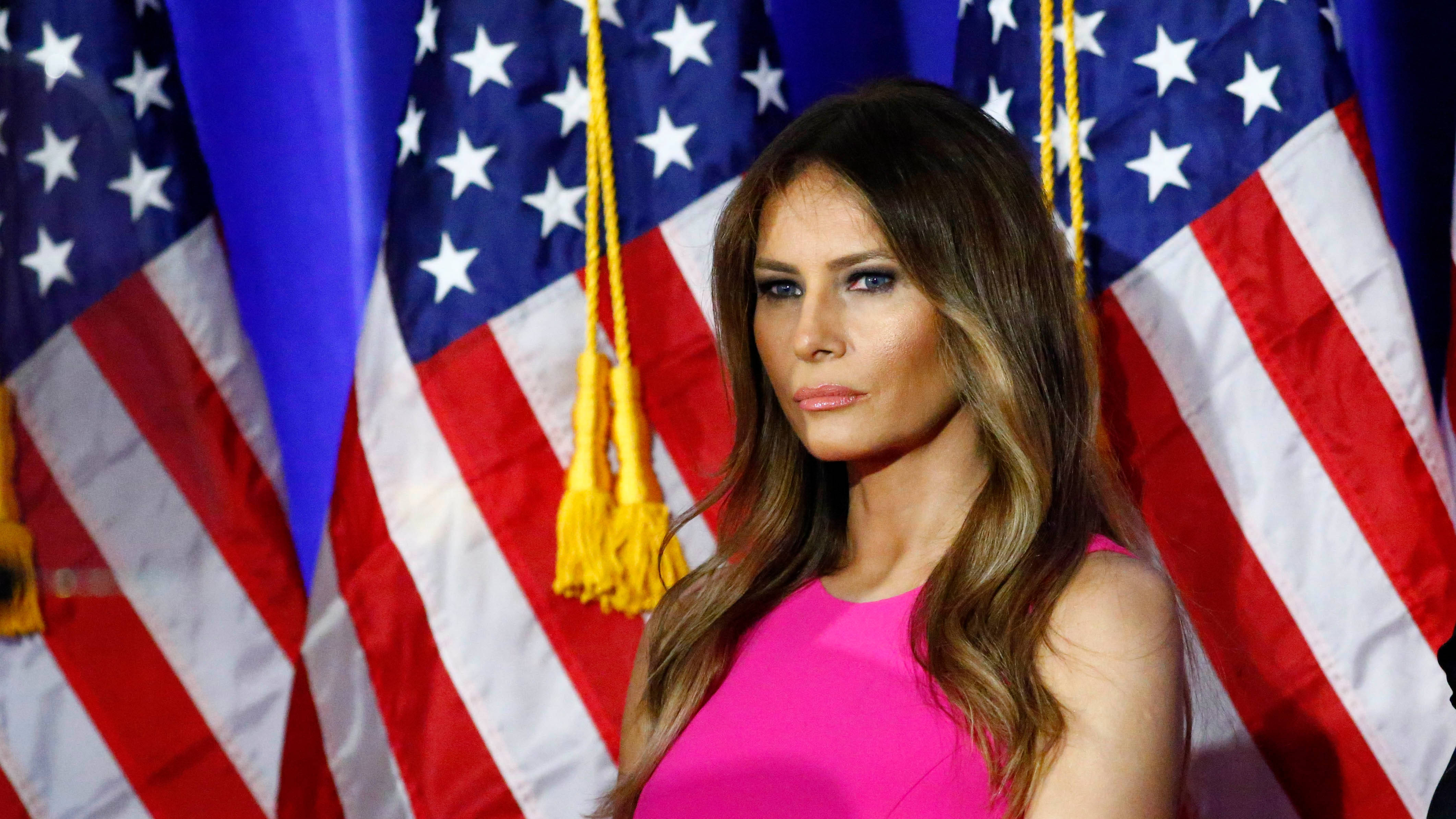 Melania 桌布 壁紙 — 梅蘭妮亞·川普身穿鮮豔粉紅色連衣裙，站在帶有金色流蘇的美國國旗背景前，姿態優雅。
