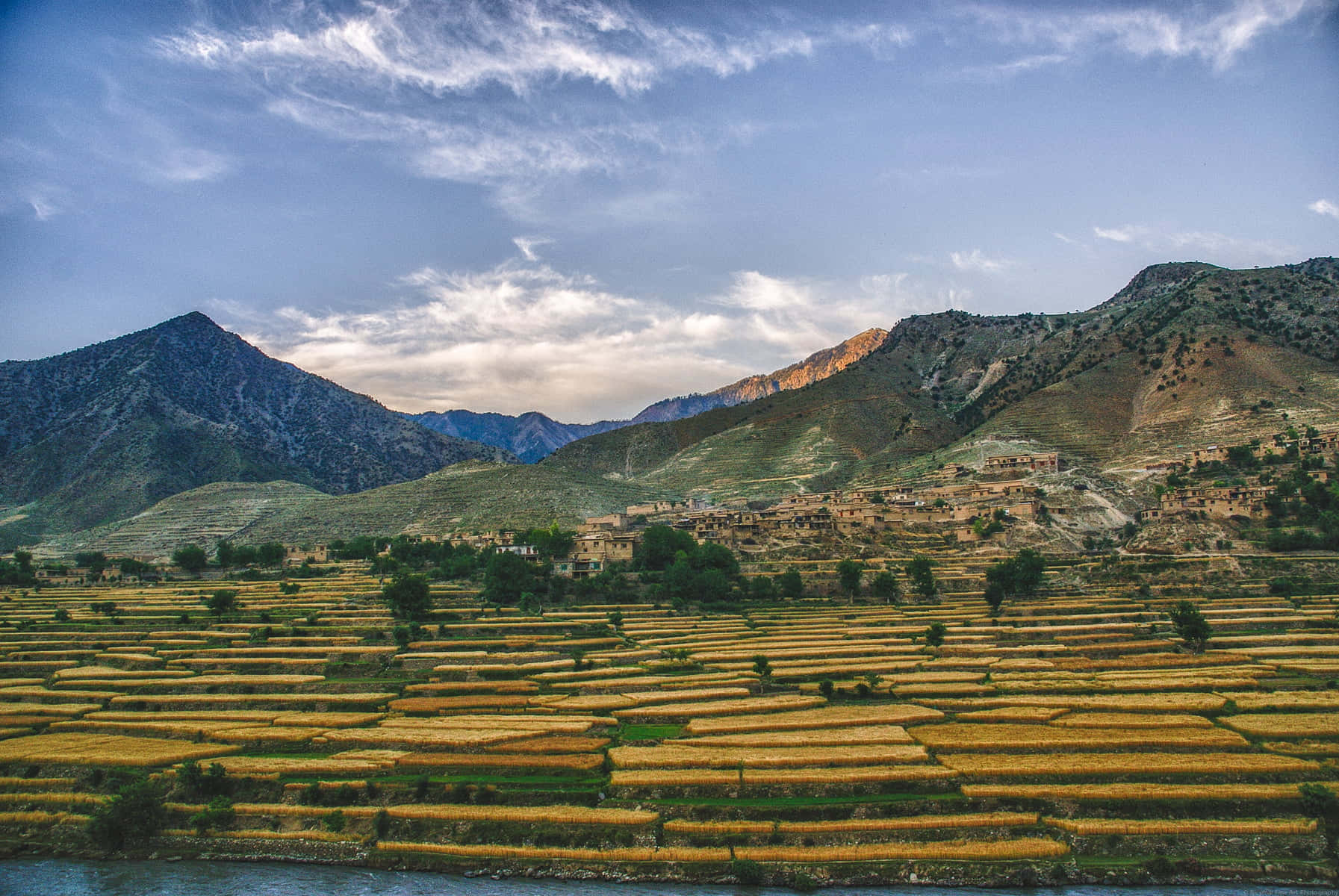 Afghanistan 桌布 壁紙 — 梯田式的金色稻田沿山坡層層疊疊流向河流，村莊坐落在群山之間，天空部分多雲。