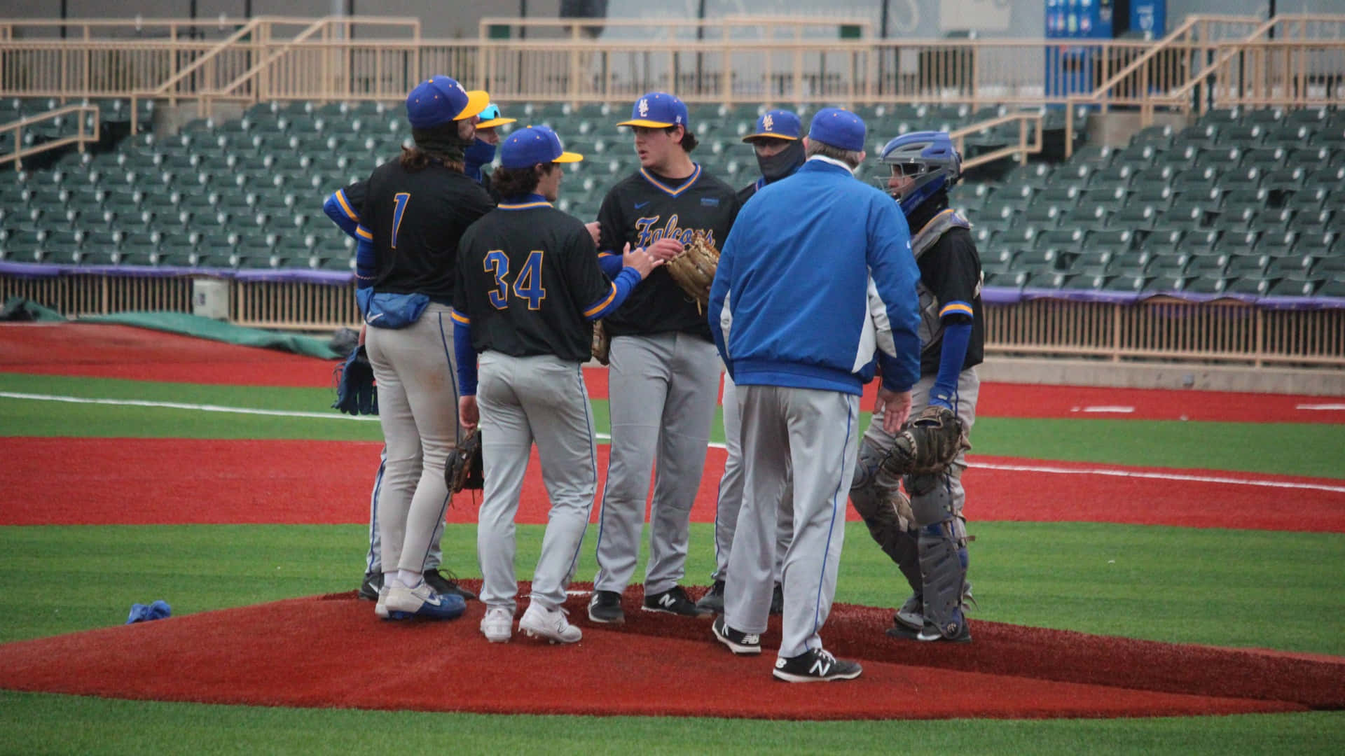 College Baseball 桌布 壁紙 — 棒球教練和球員在日間比賽中聚集在投手丘上，身後是空無一人的體育場座位。