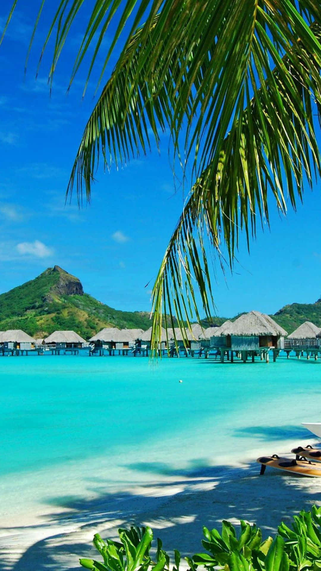 Resort 桌布 壁紙 — 棕櫚葉框住碧綠海水，水上小屋和翠綠山巒在清澈藍天下。