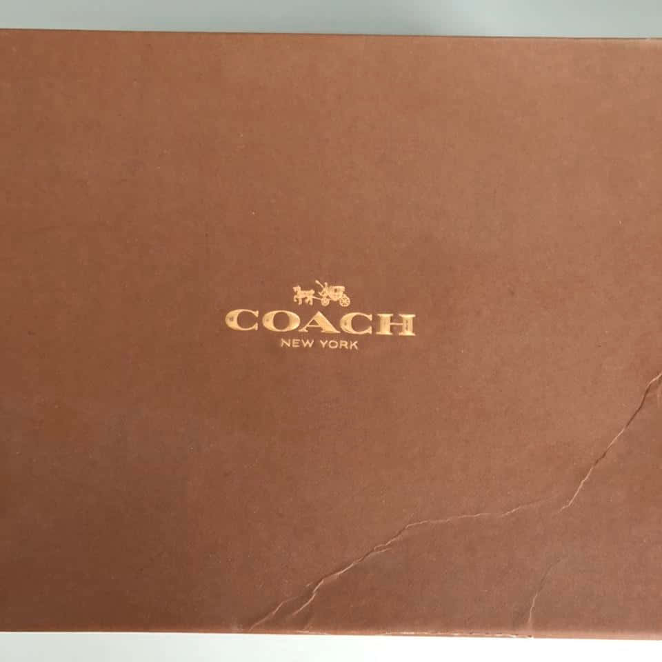教練 桌布 壁紙 — 棕色 Coach New York 品牌盒子的特寫，帶有金色箔標誌和紋理表面上可見的摺痕。