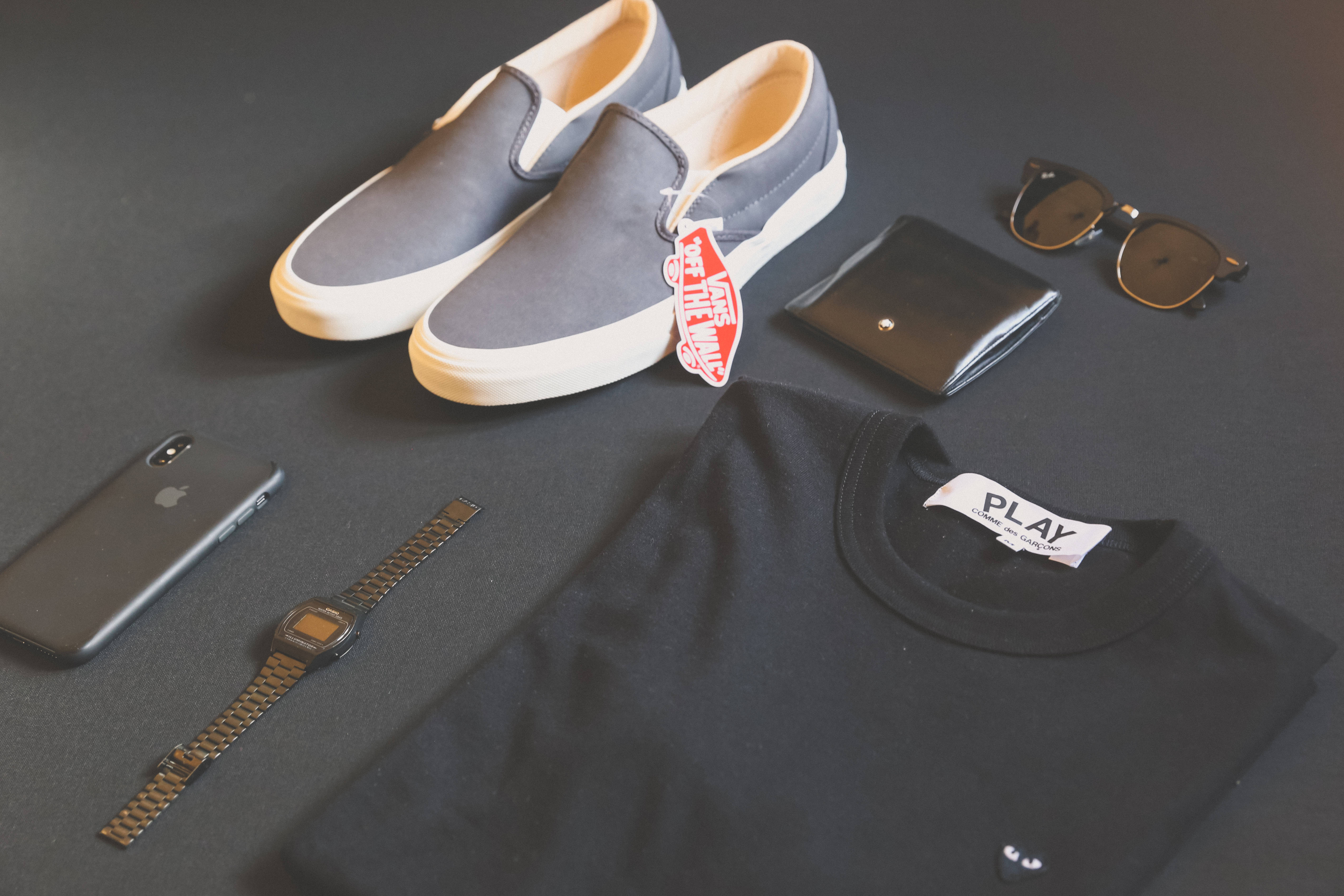 Vans 桌布 壁紙 — 極簡平鋪擺拍休閒街頭服飾單品，包括灰色懶人運動鞋、黑色T恤、手錶、太陽眼鏡和手機在深色表面上。