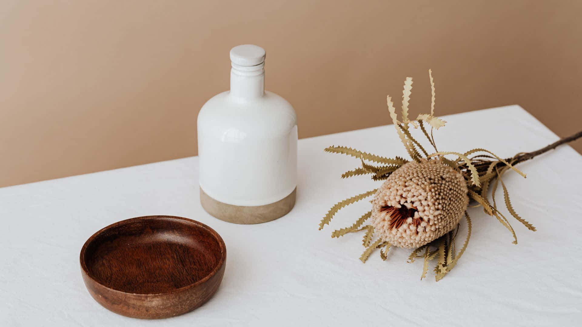 Brown Pastel Aesthetic 桌布 壁紙 — 極簡靜物，木碗、白色瓶子和乾花放在白色桌子上，背景是米色牆壁。