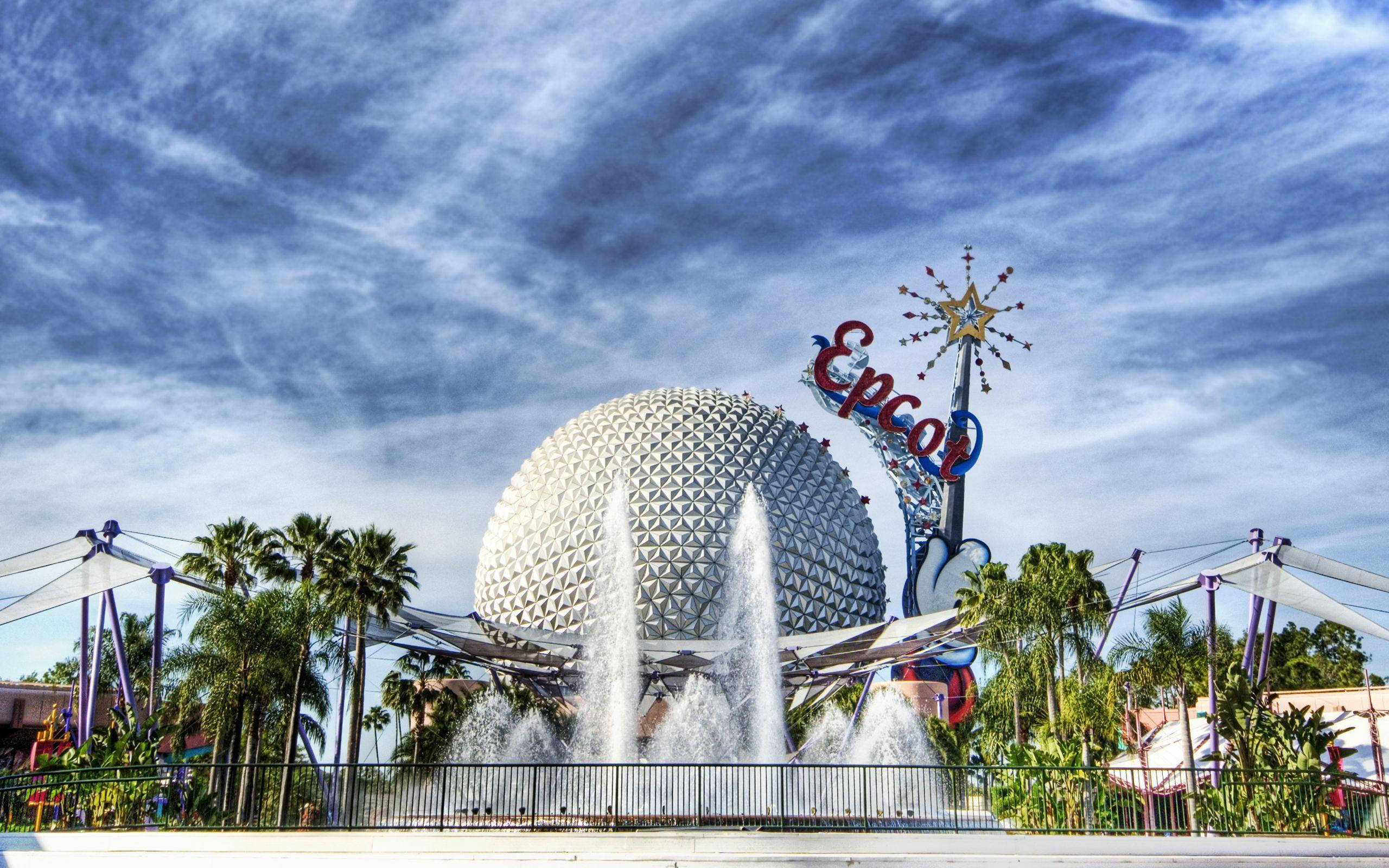 建築 桌布 壁紙 — 標誌性的 Epcot 球體在戲劇性的藍天下閃耀，噴泉在棕櫚樹和 Epcot 標誌前噴水。