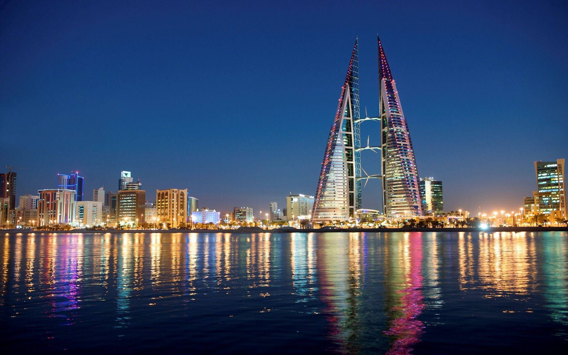 Bahrain 桌布 壁紙 — 標誌性的巴林世界貿易中心塔樓在夜間閃爍著彩色燈光，倒映在平靜的水面上。