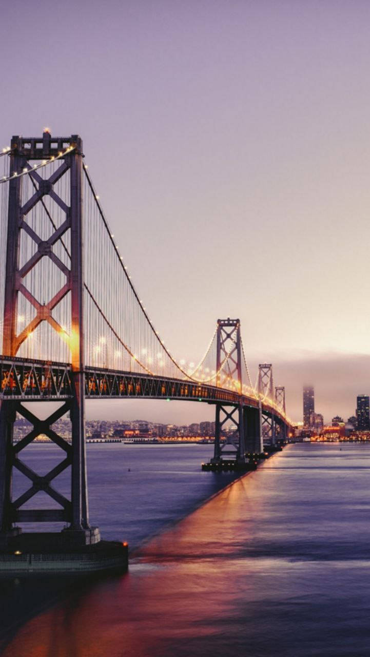san francisco iPhone 桌布 壁紙 — 標誌性的海灣大橋在暮色中溫暖地發光，其燈光倒映在下方平靜的水面上。