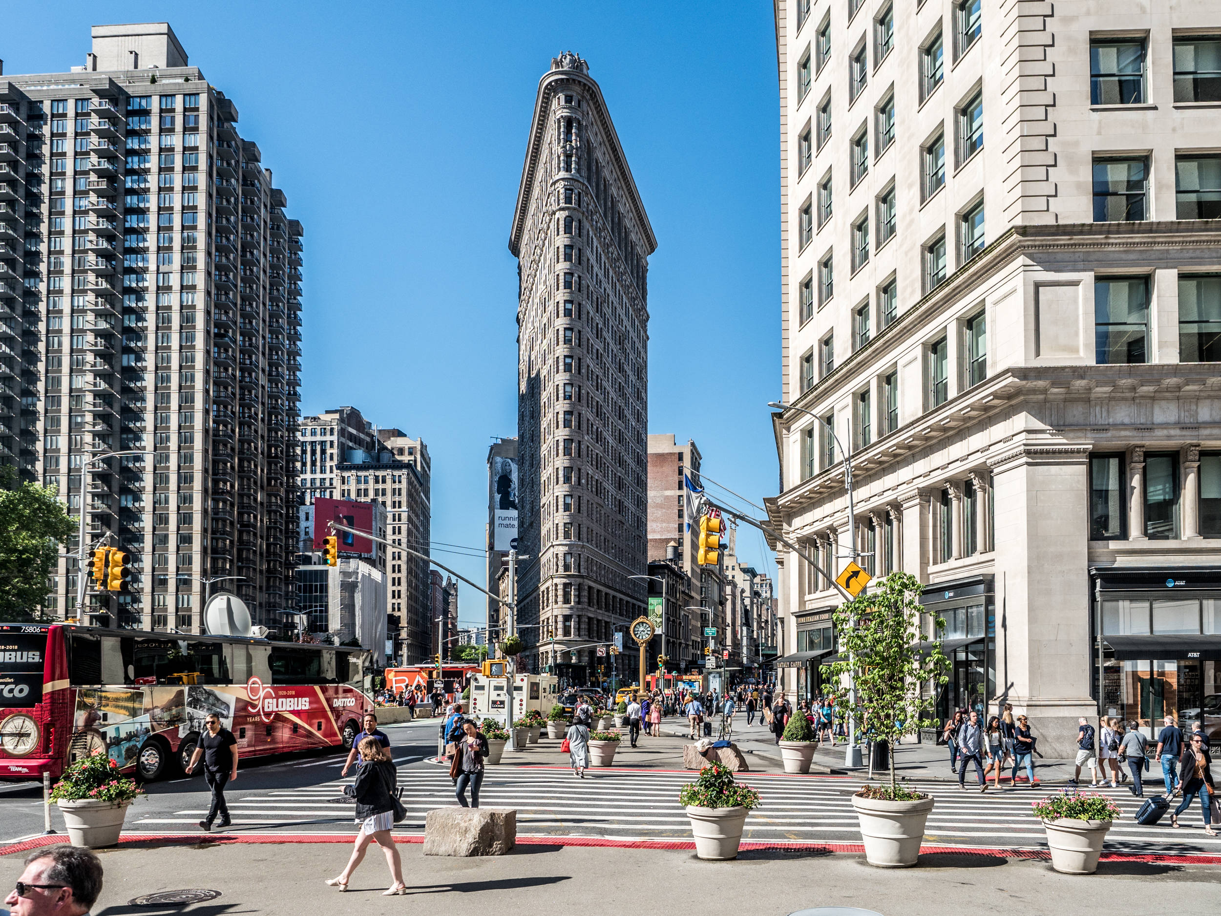 Flatiron Building 桌布 壁紙 — 標誌性的熨斗大廈在熙熙攘攘的紐約市街道上高聳入雲，街道上滿是行人和紅色雙層巴士，天空湛藍清澈。