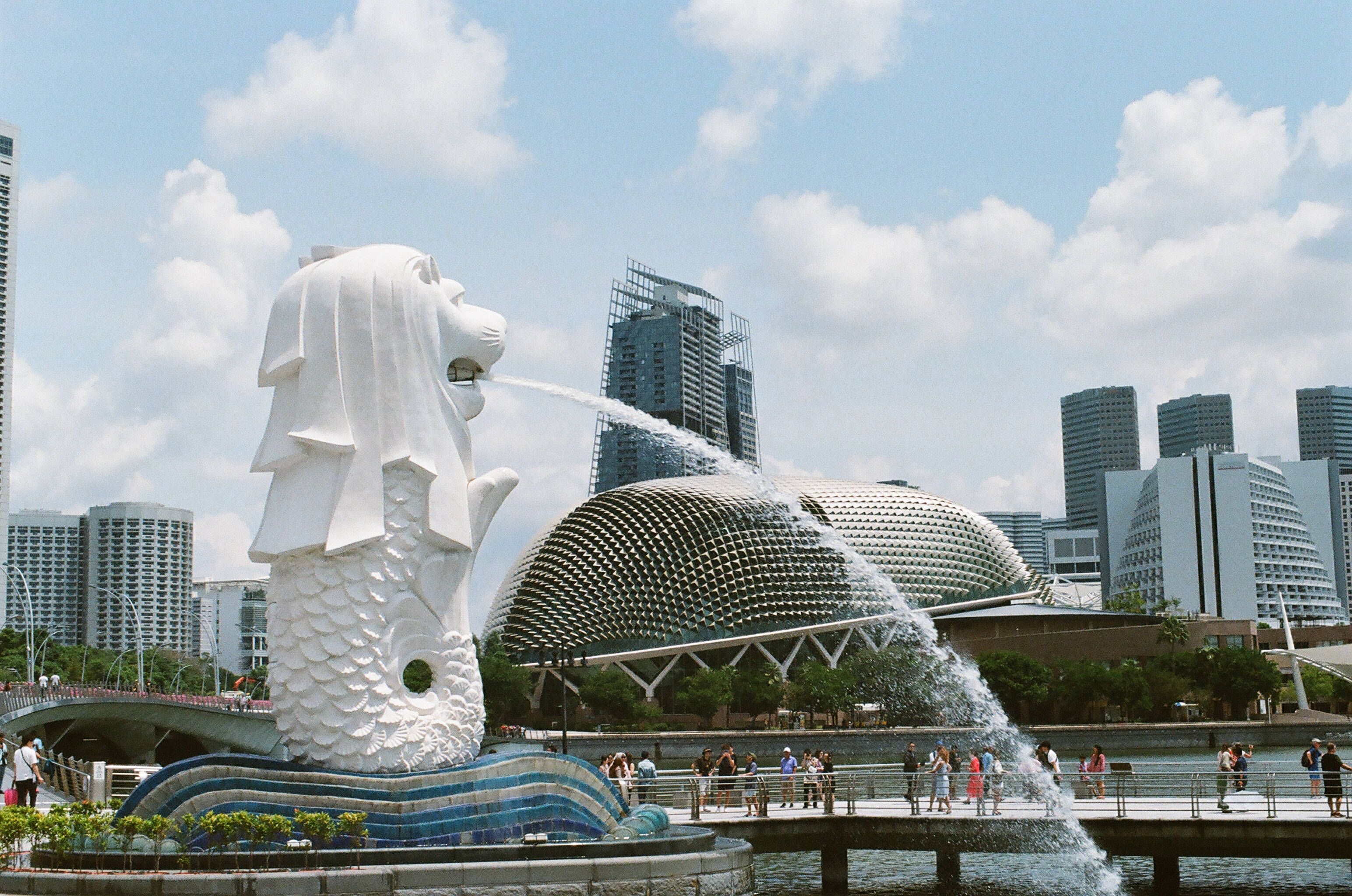 Singapore 桌布 壁紙 — 標誌性的白色魚尾獅雕像在部分多雲的天空下，以現代新加坡天際線和圓頂建築為背景，向附近有人的水池噴水。