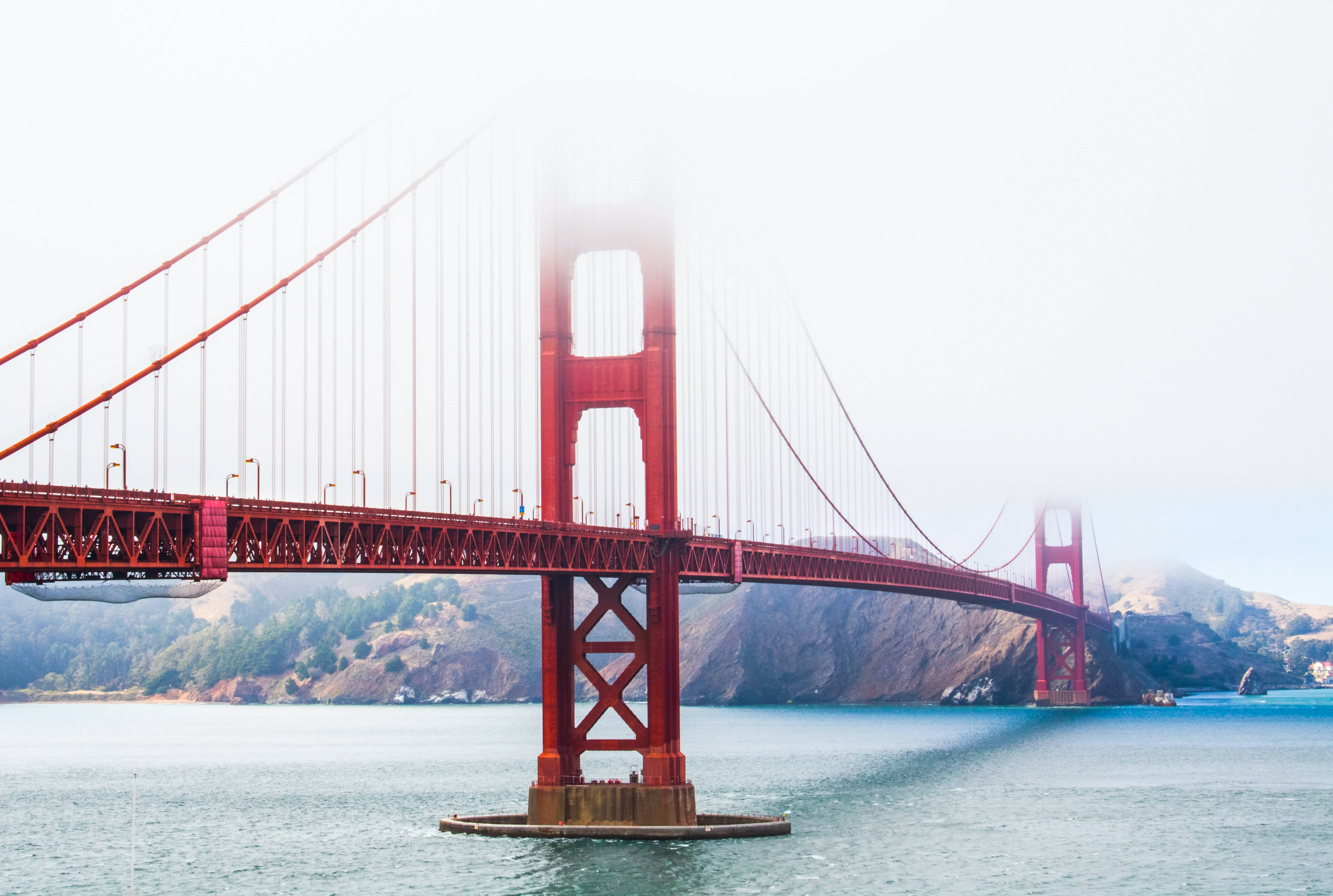 桌布 壁紙 — 標誌性的紅色金門大橋從海岸霧中浮現，下方是平靜的藍色海水，背景是朦朧的山丘。