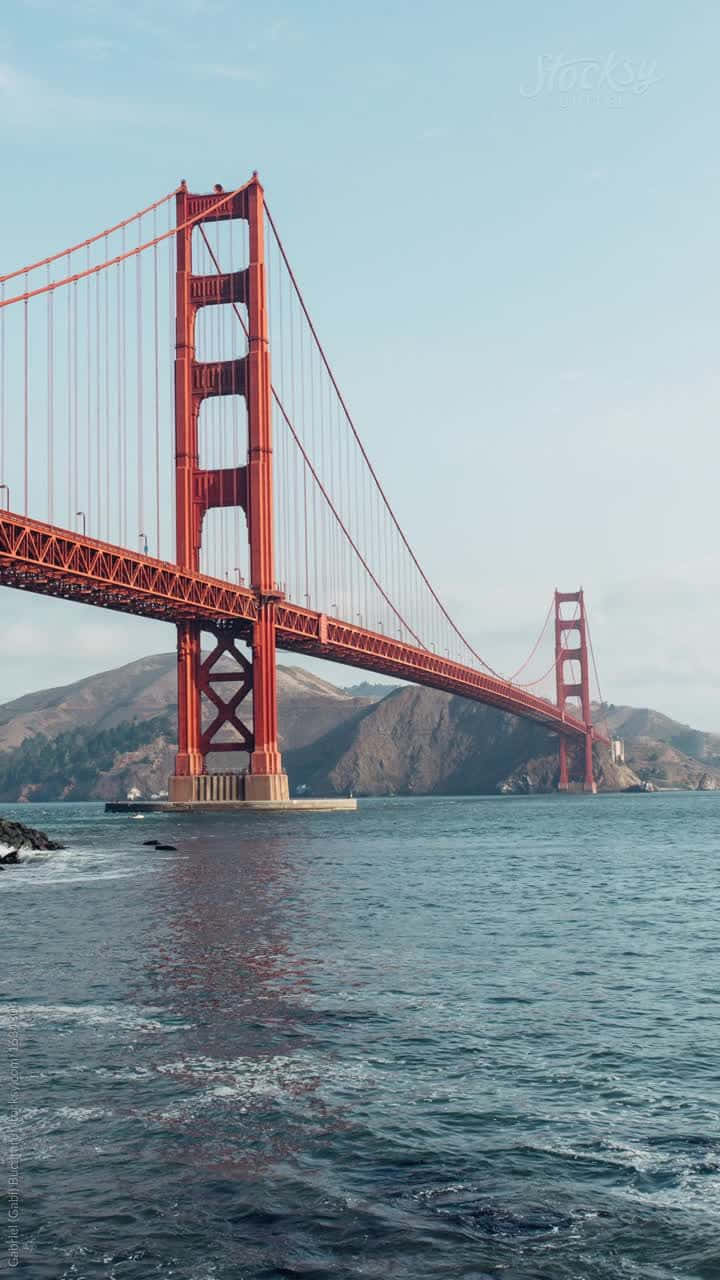 bridge 桌布 壁紙 — 標誌性的紅色金門大橋橫跨在平靜的藍色海灣上，背景是朦朧的山丘，天空晴朗。