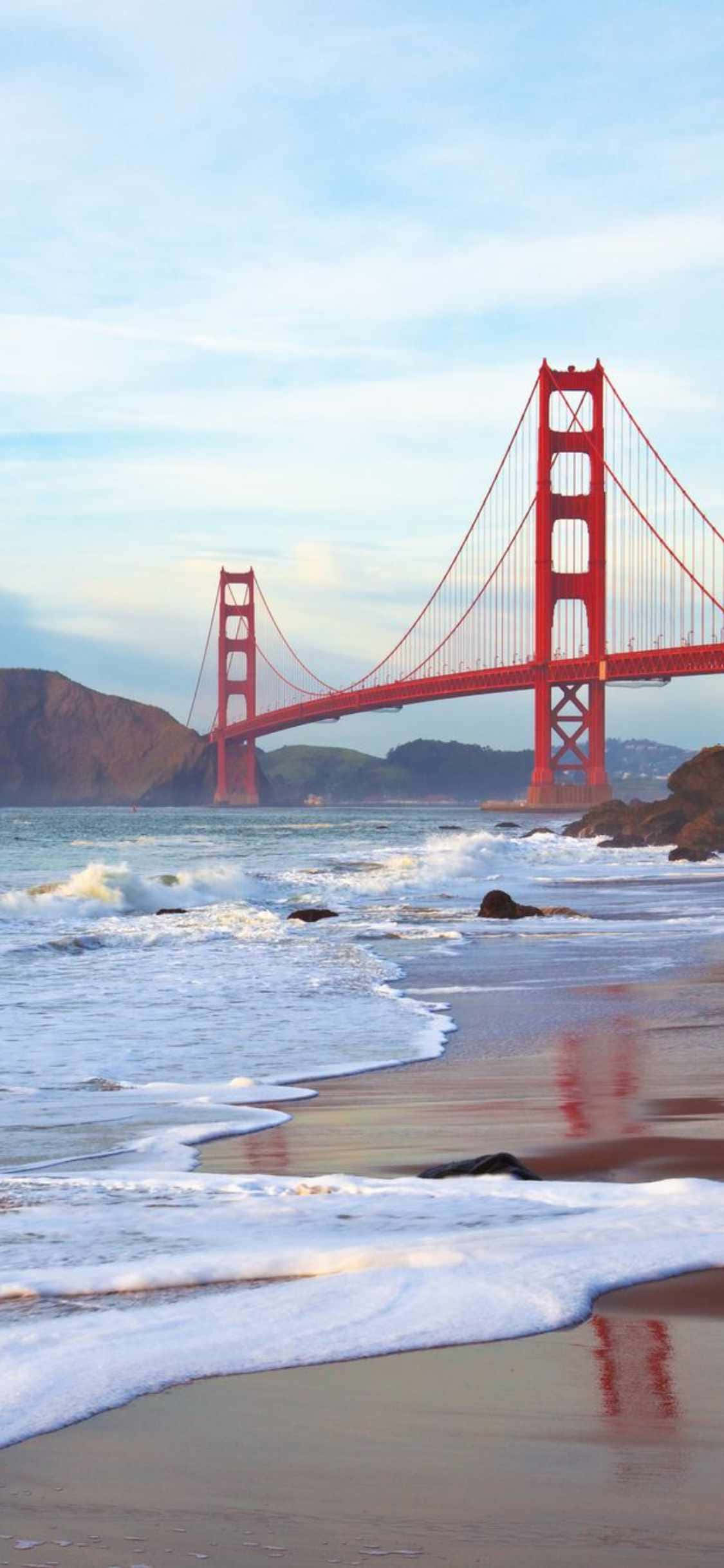 iPhone Xs Malibu 桌布 壁紙 — 標誌性的紅色金門大橋橫跨海灣，海浪拍打，沙灘在柔和的藍天下。