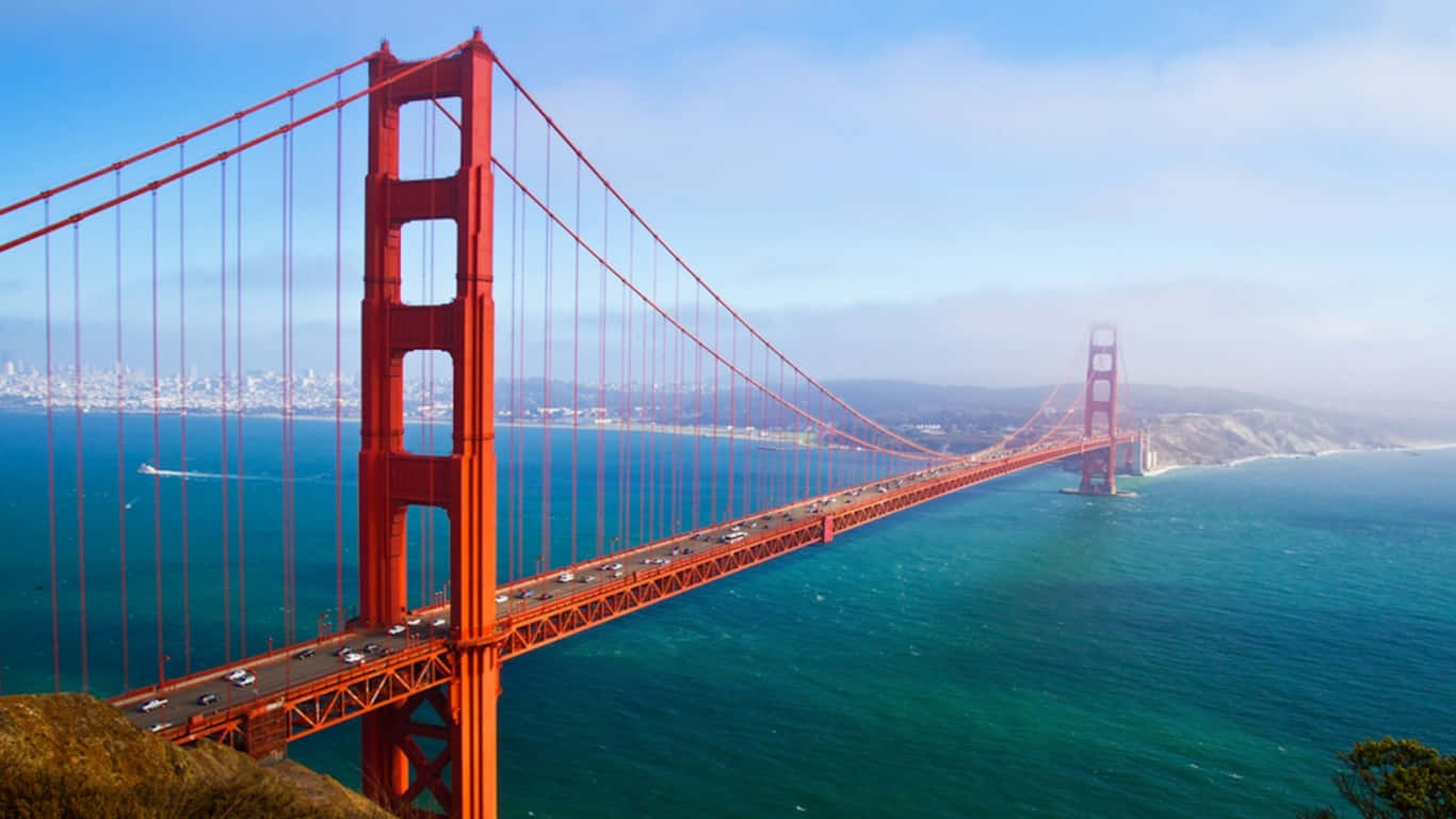 1366x768 San Francisco 桌布 壁紙 — 標誌性的紅色金門大橋橫跨深藍色的海灣，天空部分多雲，遠處有城市景觀。