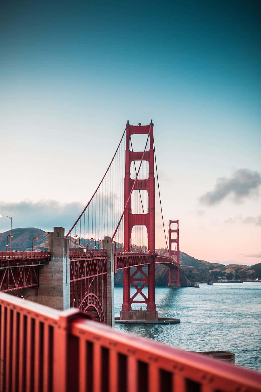 California Aesthetic 桌布 壁紙 — 標誌性的紅色金門大橋雄偉地矗立在平靜的藍色水域上，在柔和的暮色天空下，遠處有山丘。