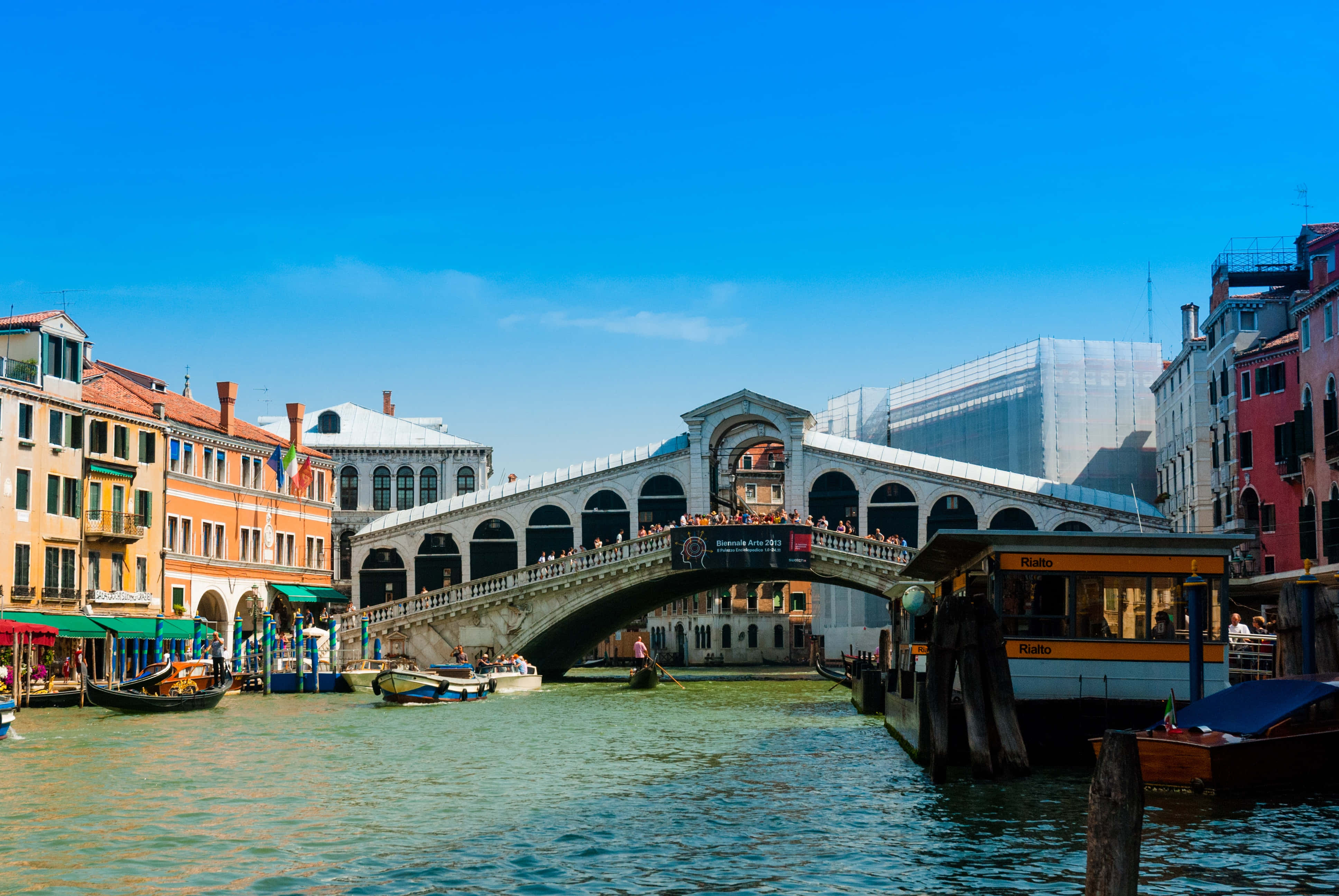 venice 桌布 壁紙 — 標誌性的里亞爾托橋在晴朗的藍天下橫跨繁忙的威尼斯運河，色彩繽紛的歷史建築沿水而立。