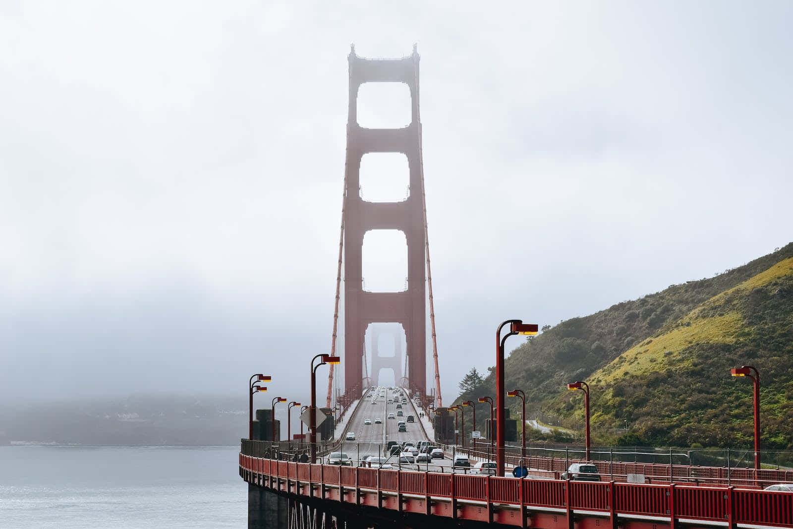 San Francisco Fog 桌布 壁紙 — 標誌性的金門大橋在霧天中高聳，汽車沿著其紅色車道行駛，旁邊是朦朧的海灣。