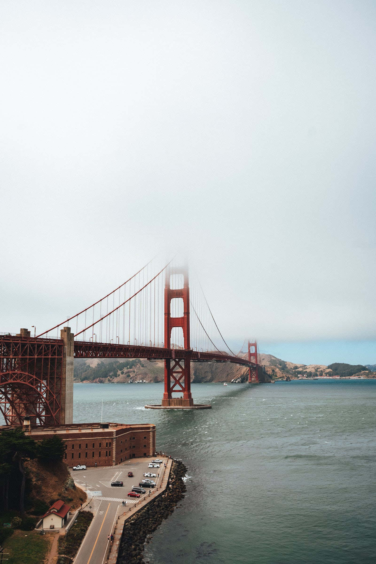 San Francisco Fog 桌布 壁紙 — 標誌性的金門大橋在霧濛濛的灰色天空下巍然聳立，其紅橙色結構橫跨平靜的海灣，遠處有山丘。