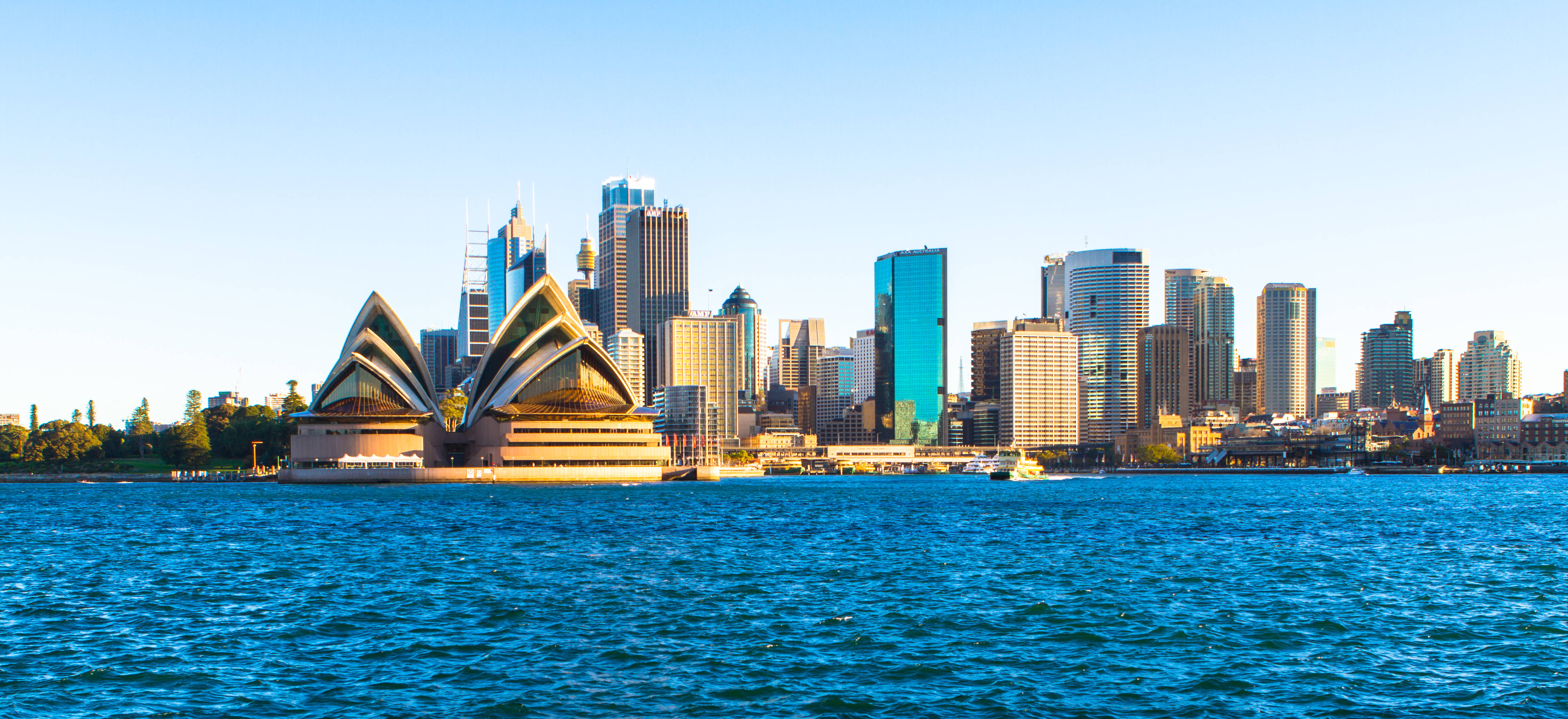 Sydney 桌布 壁紙 — 標誌性的雪梨歌劇院在清澈的藍天下閃耀，旁邊是波光粼粼的港灣水域，背景是現代化的城市天際線。