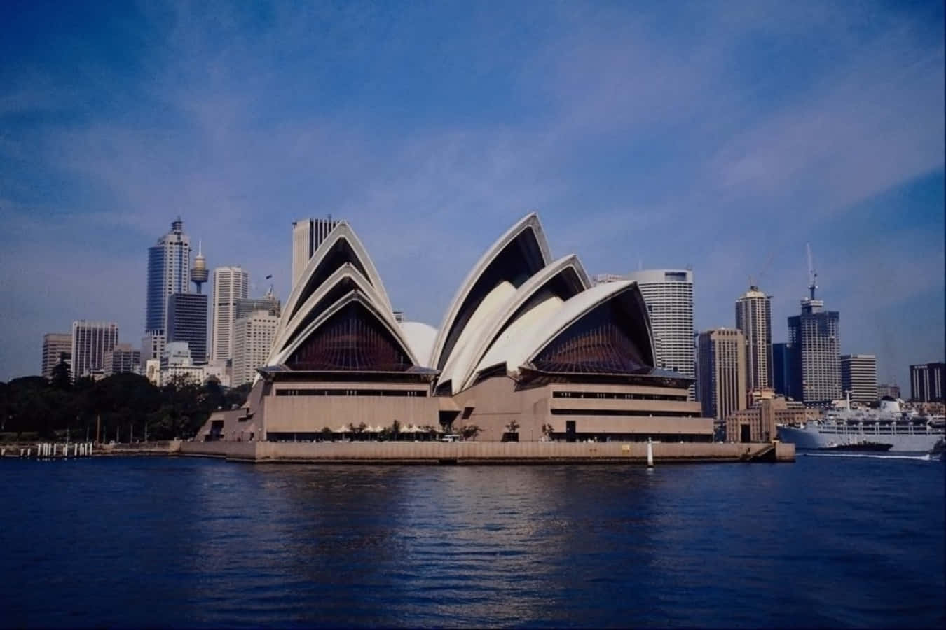 建築 桌布 壁紙 — 標誌性的雪梨歌劇院擁有帆狀屋頂，在清澈的藍天和背景中的城市摩天大樓映襯下，雄偉地矗立在水面上。