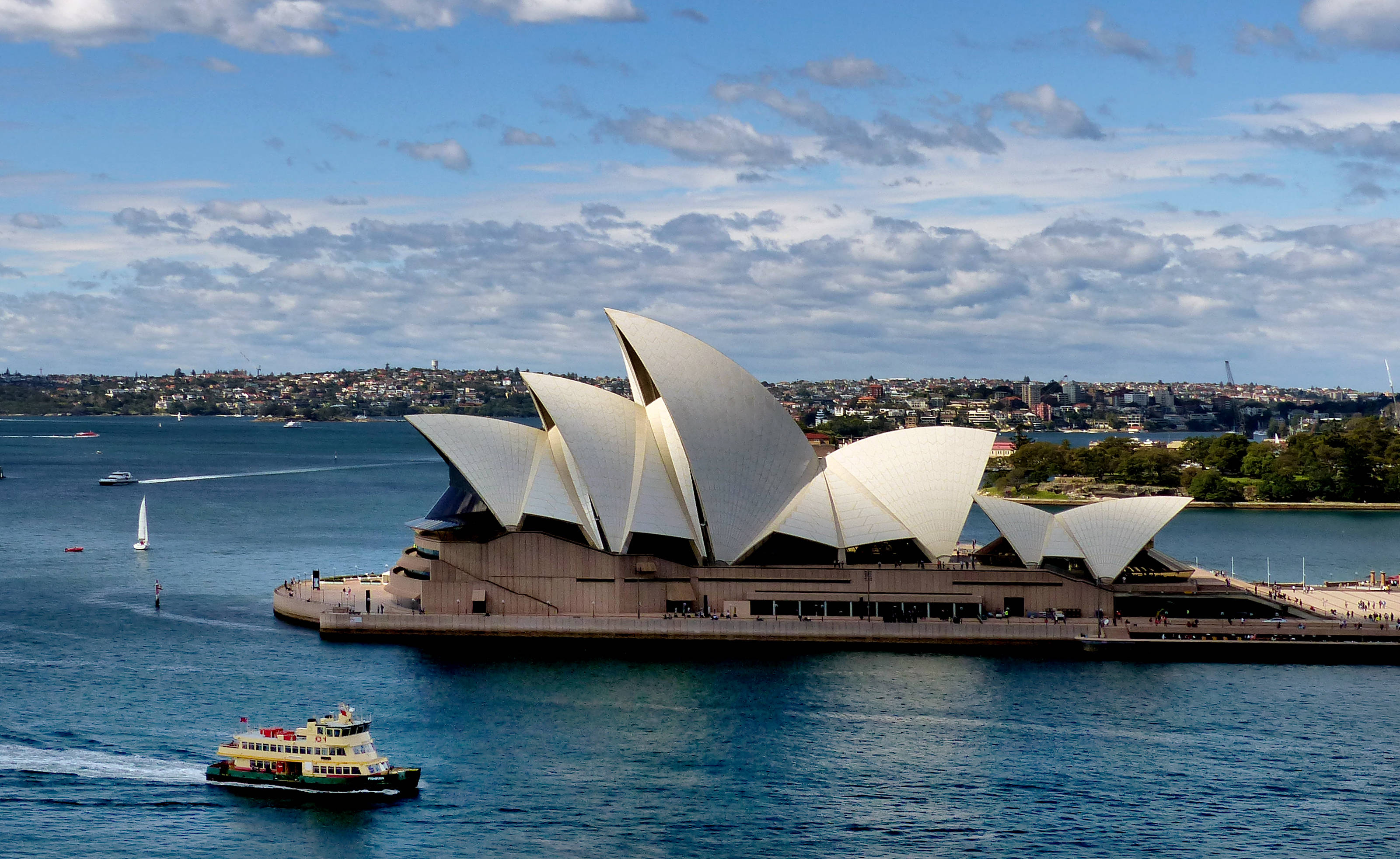 Sydney 桌布 壁紙 — 標誌性的雪梨歌劇院雄偉地矗立在港灣上，帆船和渡輪在部分多雲的天空下於藍色水面上滑行。